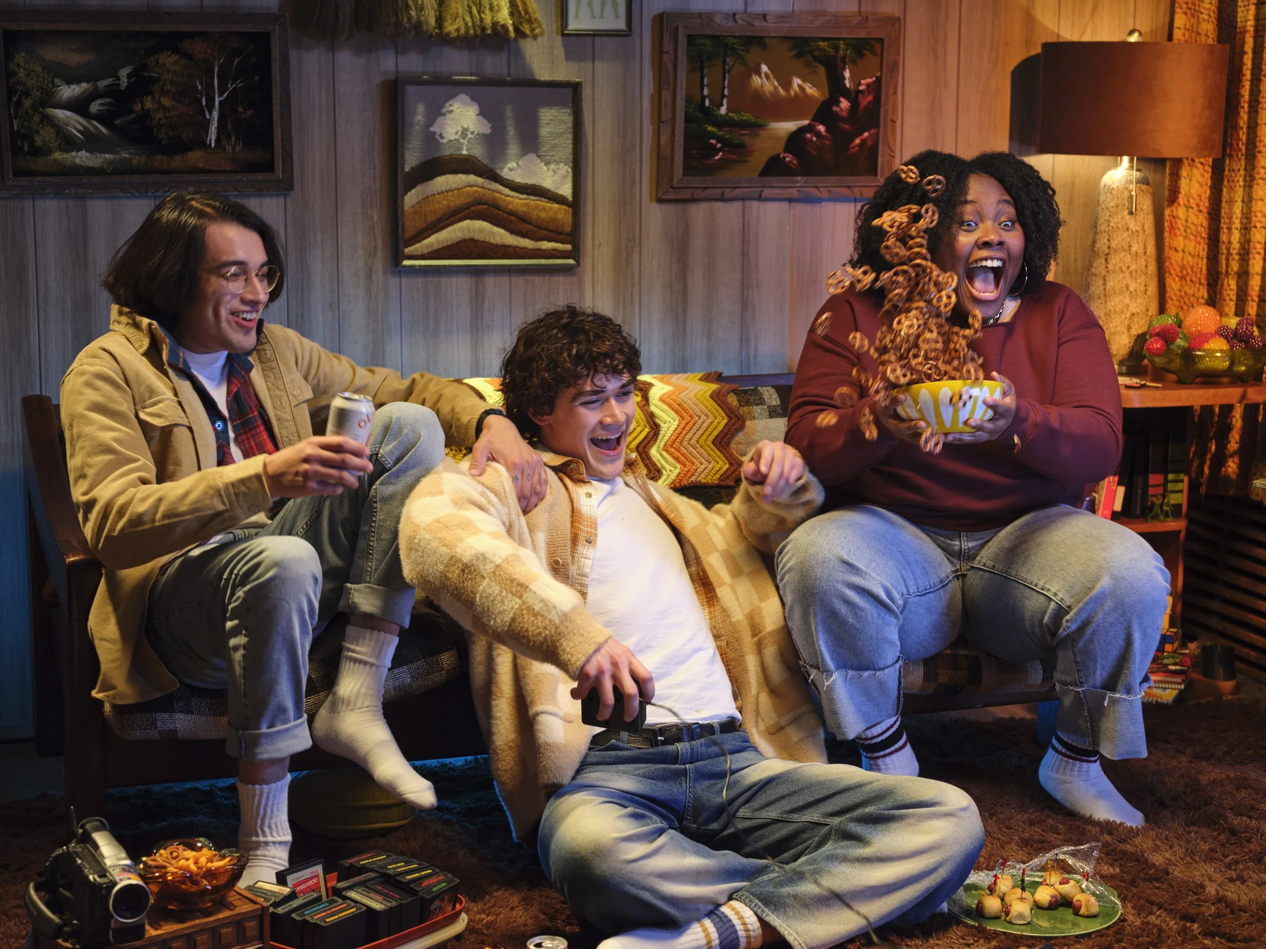 Three friends are sitting on a retro-style living room couch, laughing and having fun as they watch a movie; one woman is eating popcorn from a yellow bowl, another man sitting on the floor is holding a game controller, and a third man is holding a s