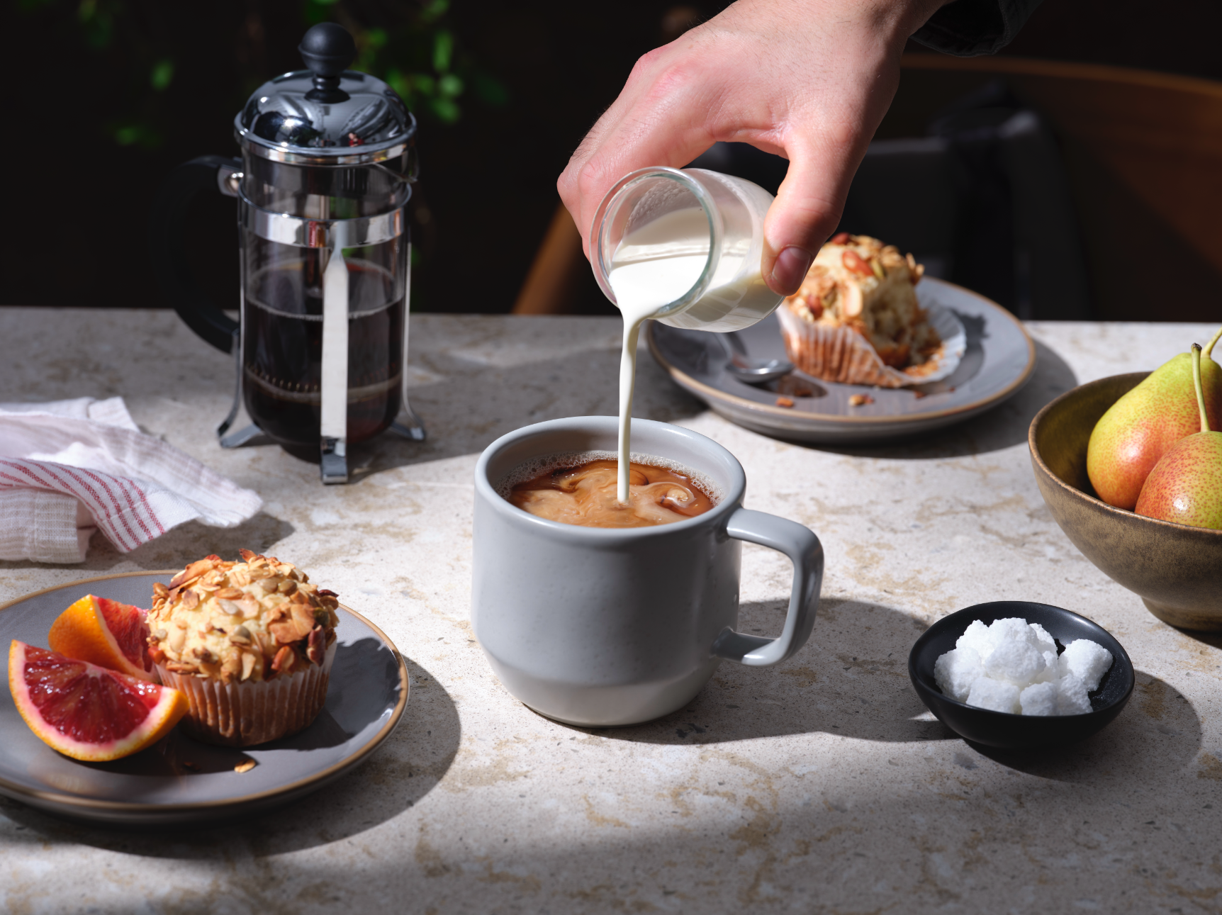 a hand pouring cream into a coffee mug surrounded by fresh fruit, muffins and coffee. 
