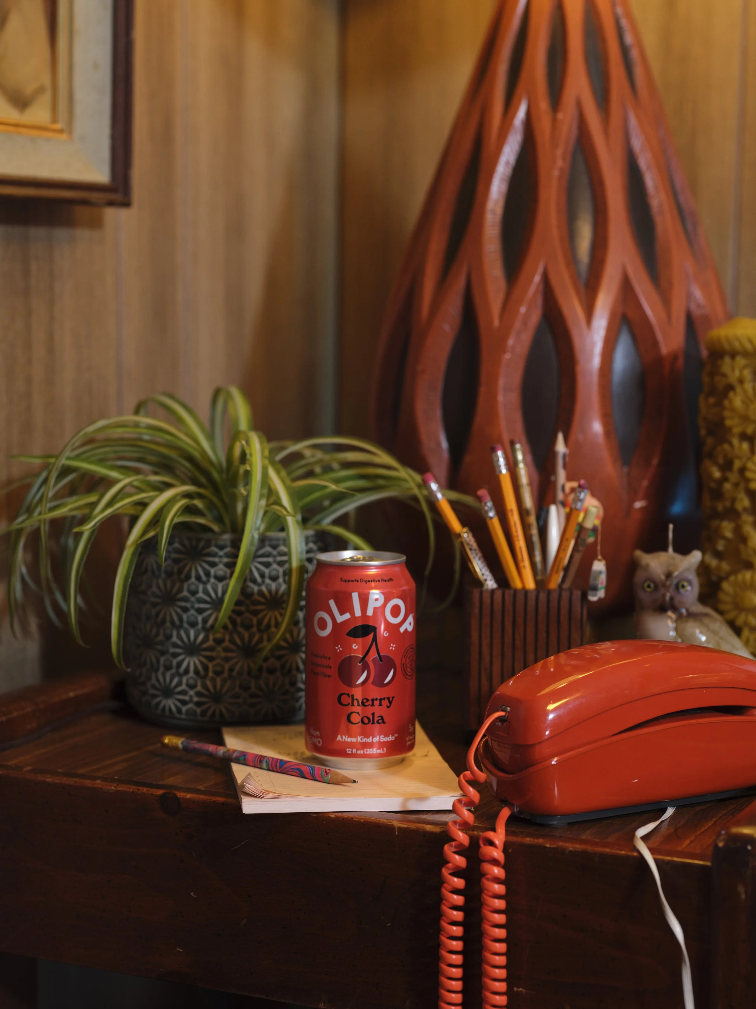 A wooden desk with a potted plant, a red can labeled 'Olipop Cherry Cola' on top of a notepad, a colorful pencil, a red corded telephone, a wooden container with pencils, and various decorative items in the background.