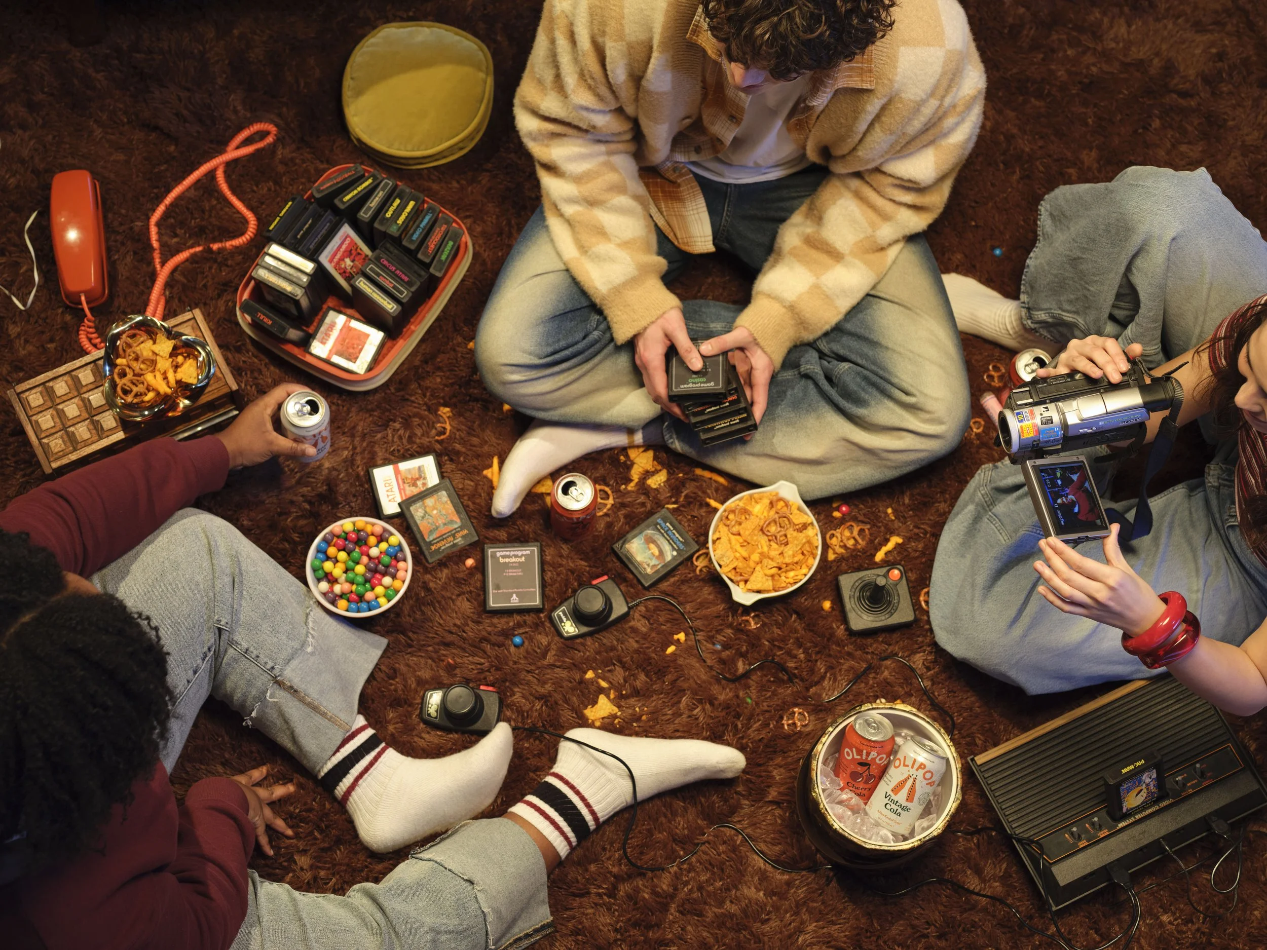 overhear shot of 3 people sitting on a shag carpet, with snacks. soda and video games on the rug