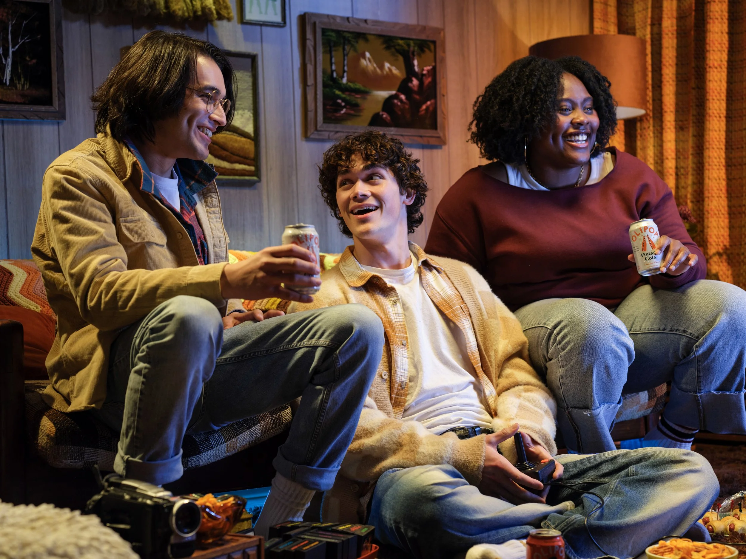 Three people sitting in a vintage 80's game room, hanging out playing video games, and drinking sodas.
