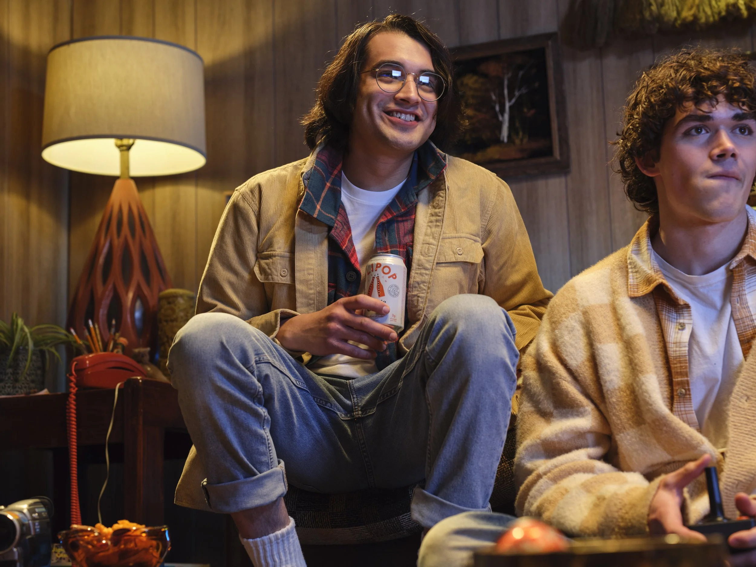 Two young men sitting in a cozy wooden room, one smiling holding a can, the other focused on a video game controller.
