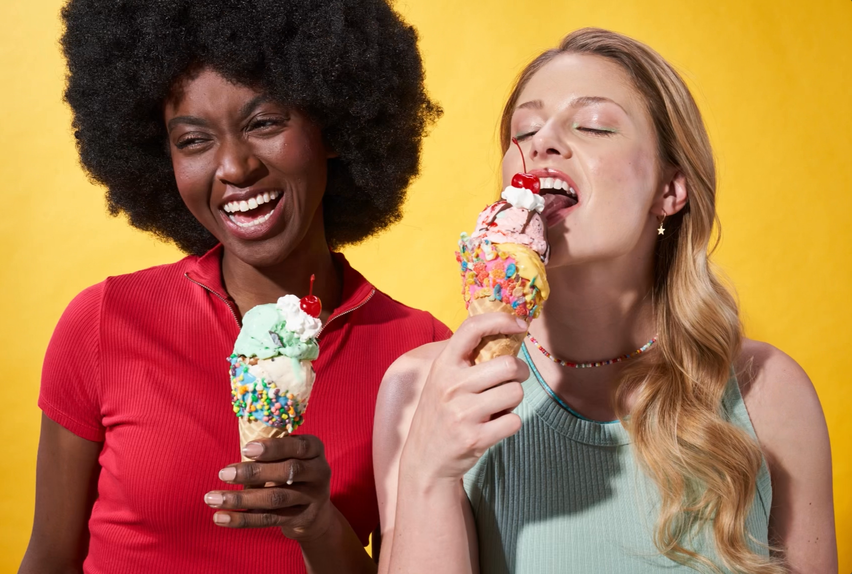 two women waiting ice cream cones smiling on a yellow background. 
