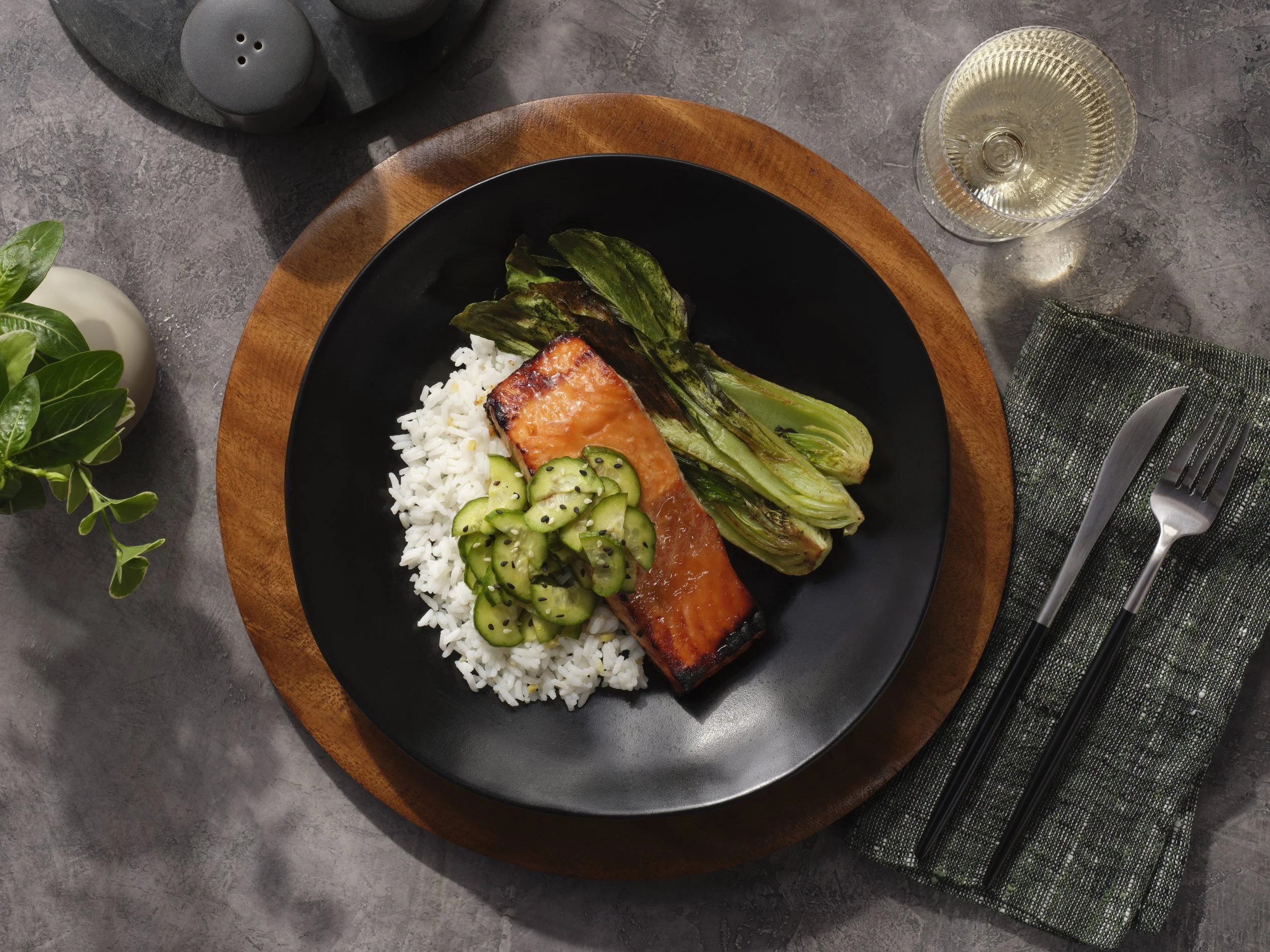 A black plate with grilled salmon, white rice, cucumber salad, and grilled bok choy on a wooden charger plate. A glass of white wine, a knife and fork on a green napkin, and a small potted plant are also visible on a gray table.