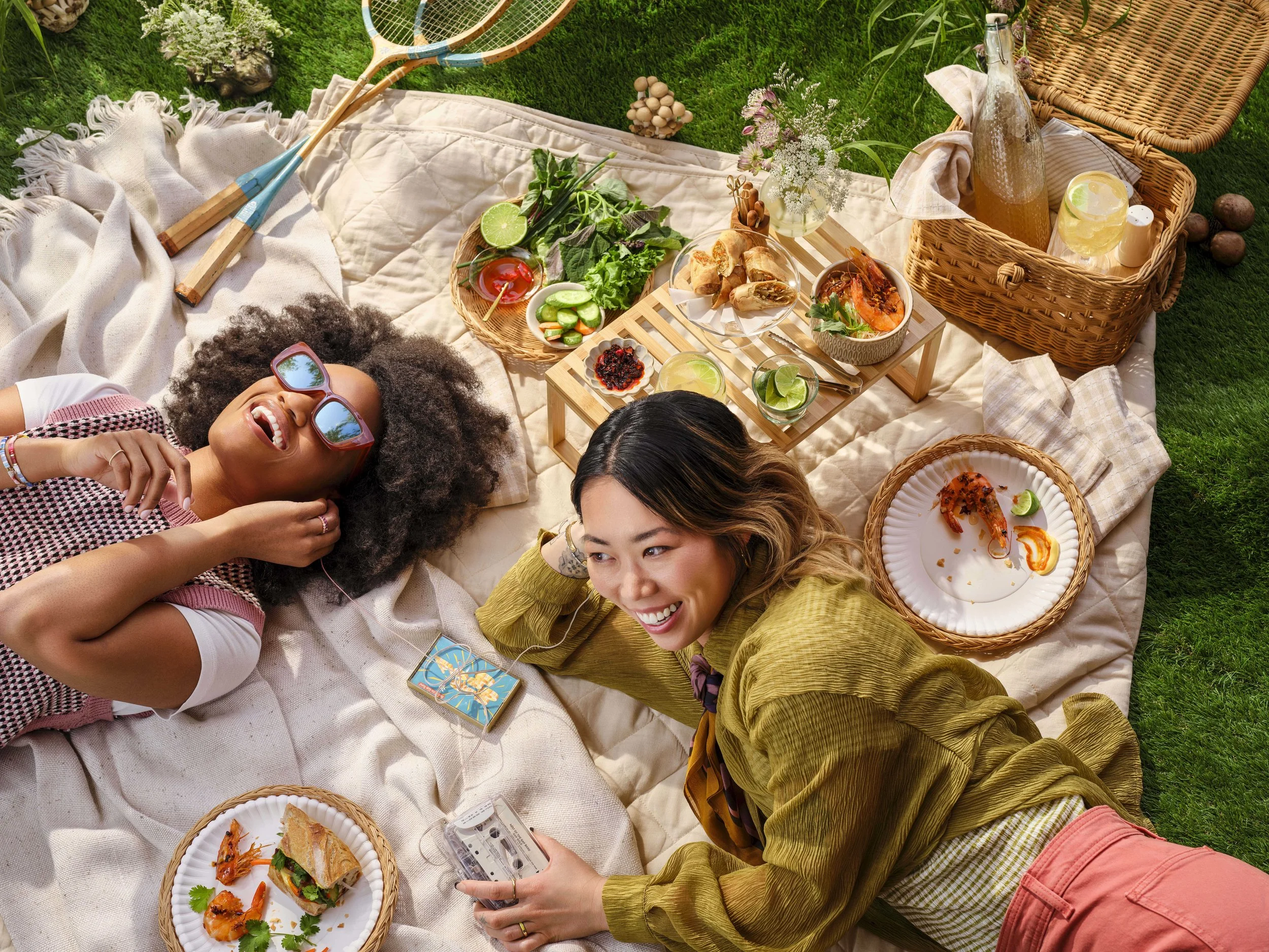 Two women laying on a picnic blanket on the grass during summer. Listening to a cassette tape and sharing headphones. Surrounded by grilled shrimp, bahn mi sandwiches and cold drinks. 
