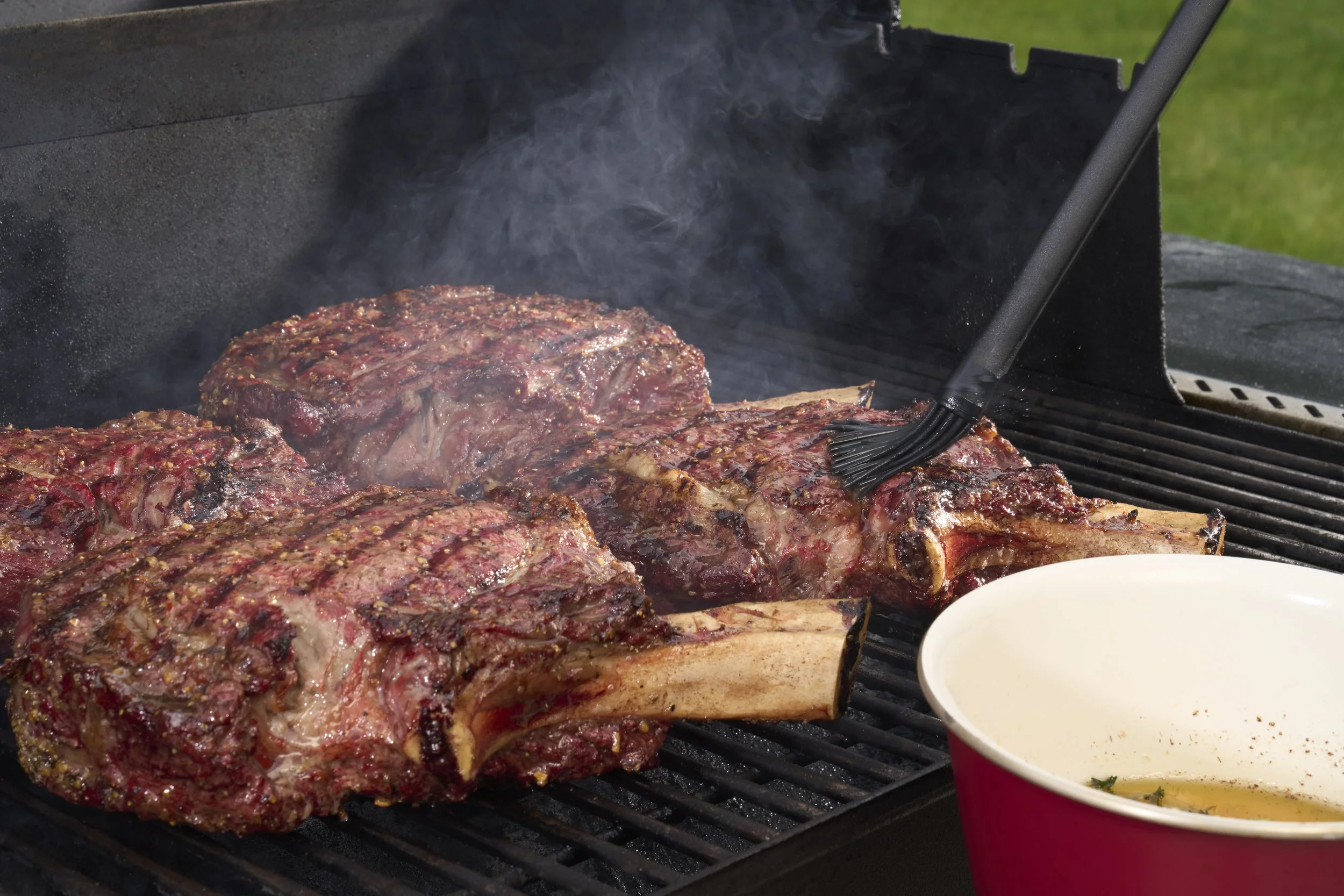 Three large, seasoned steaks with bones grilling on a barbecue, with smoke rising, while someone brushes marinade or sauce on them with a basting brush. There is a white bowl with some liquid and herbs in the foreground, and green grass in the backgr