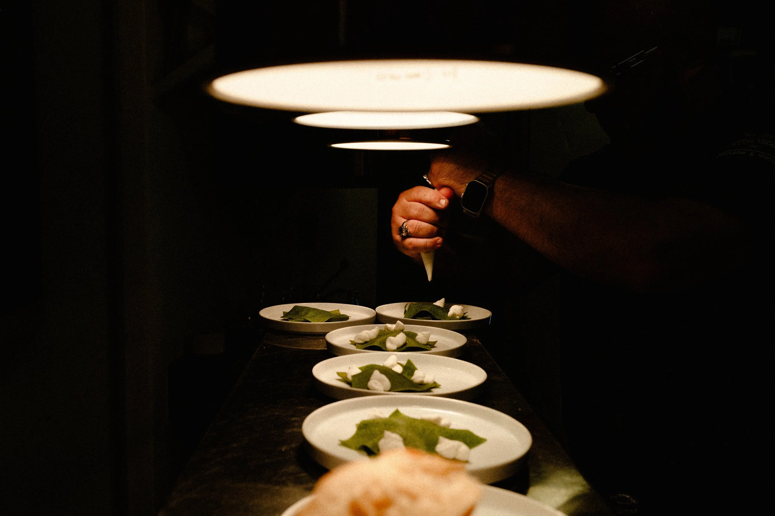 A chef decorates plates with small green leaves and white dollops of sauce under dim lighting.