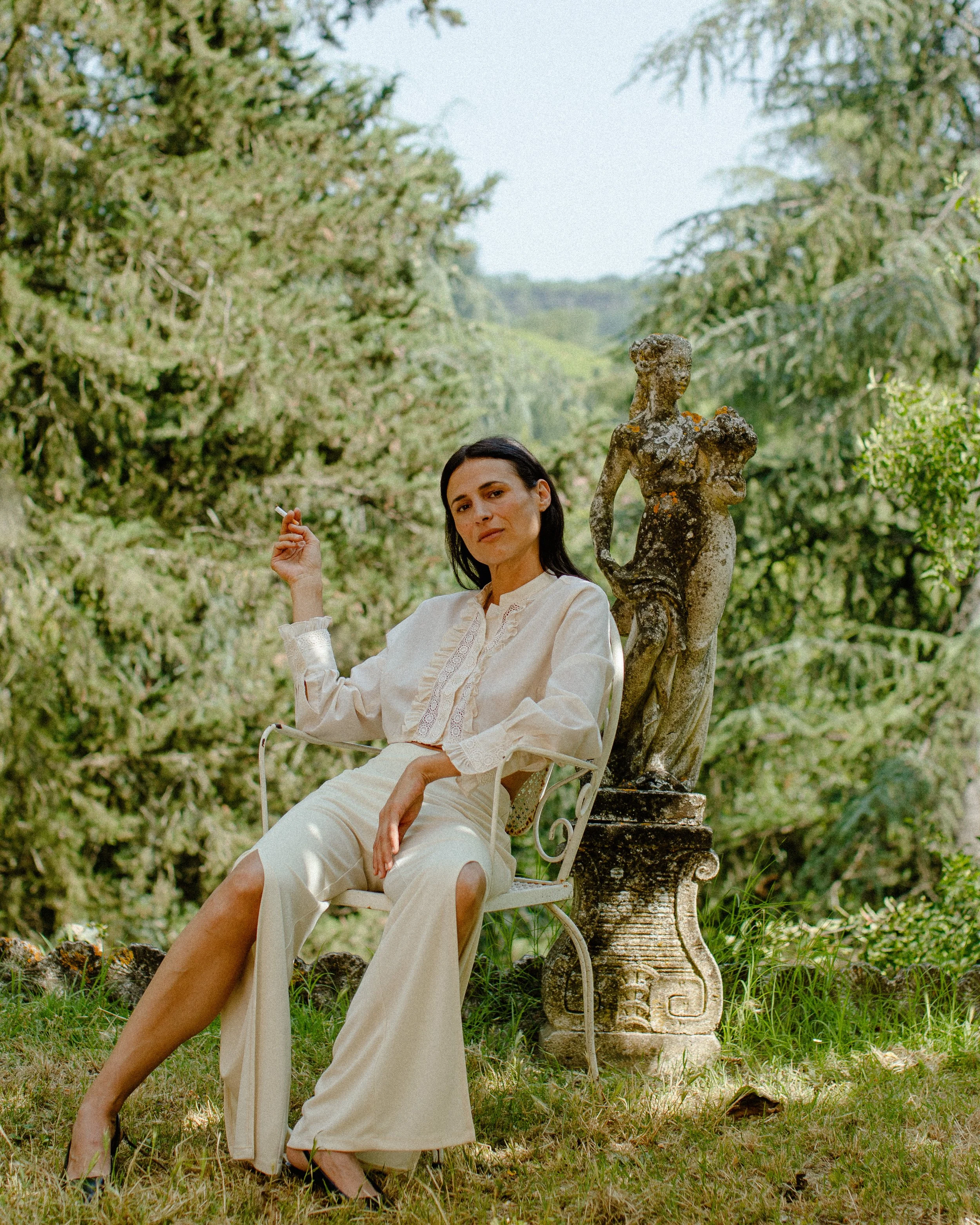 A woman sitting on a white metal chair outdoors, wearing a cream-colored outfit, with a stone sculpture of a woman beside her, surrounded by lush green trees and bushes in the background.