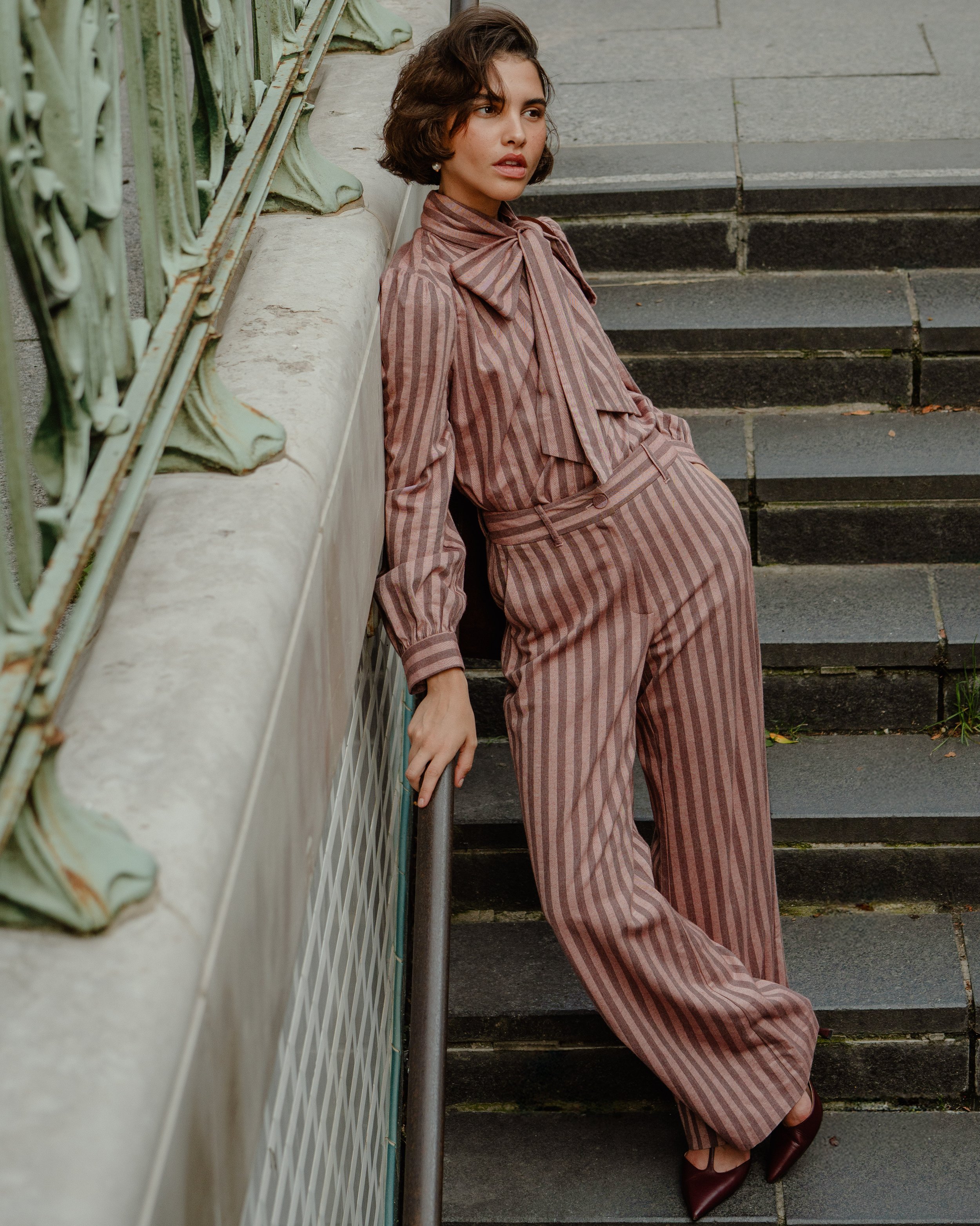 A woman with short curly brown hair wearing a pink and brown striped suit with a bow tie, leaning against a stone wall on a staircase, holding a railing.