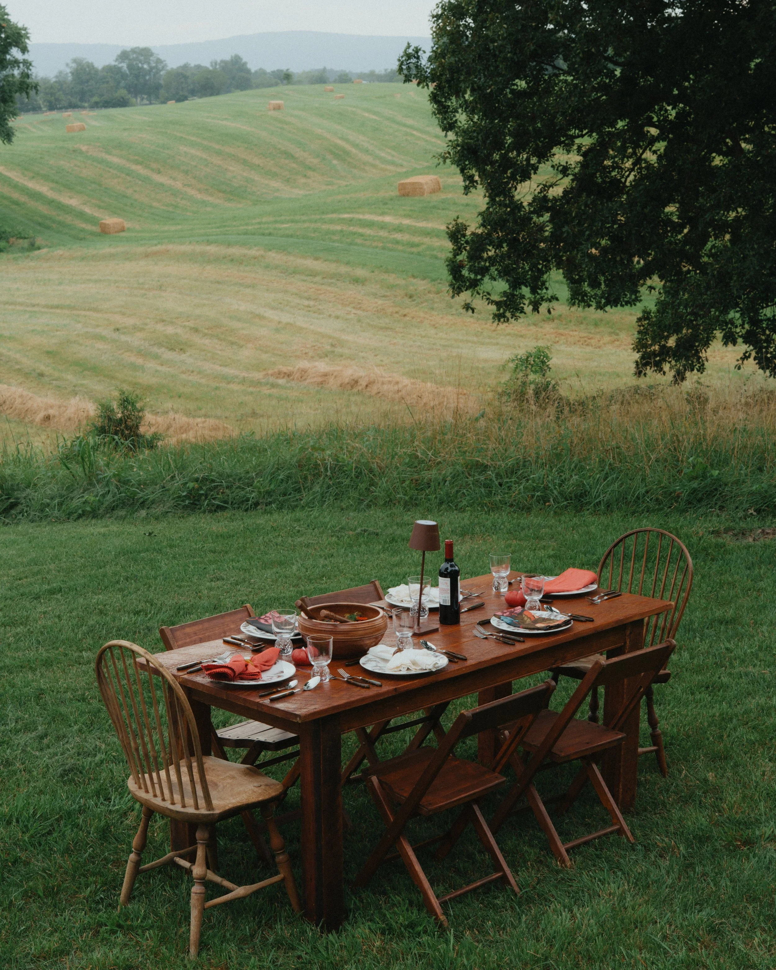 Outdoors dining table set with plates, glasses, utensils, a bottle of wine, and a lamp, on a grassy lawn with a rolling green hillside and hay bales in the background.