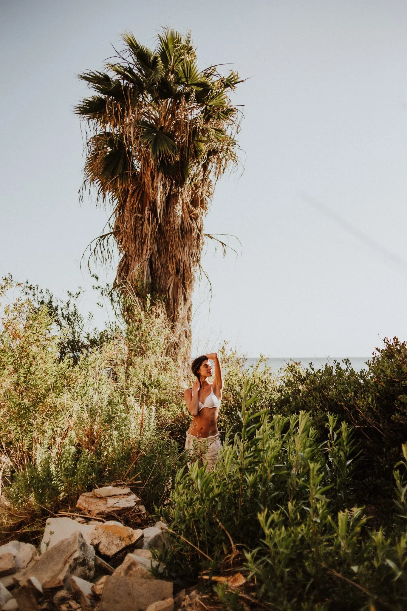 A woman in a white bikini top and light-colored shorts stands among green bushes and rocks with a tall palm tree behind her, overlooking a beach and sky.
