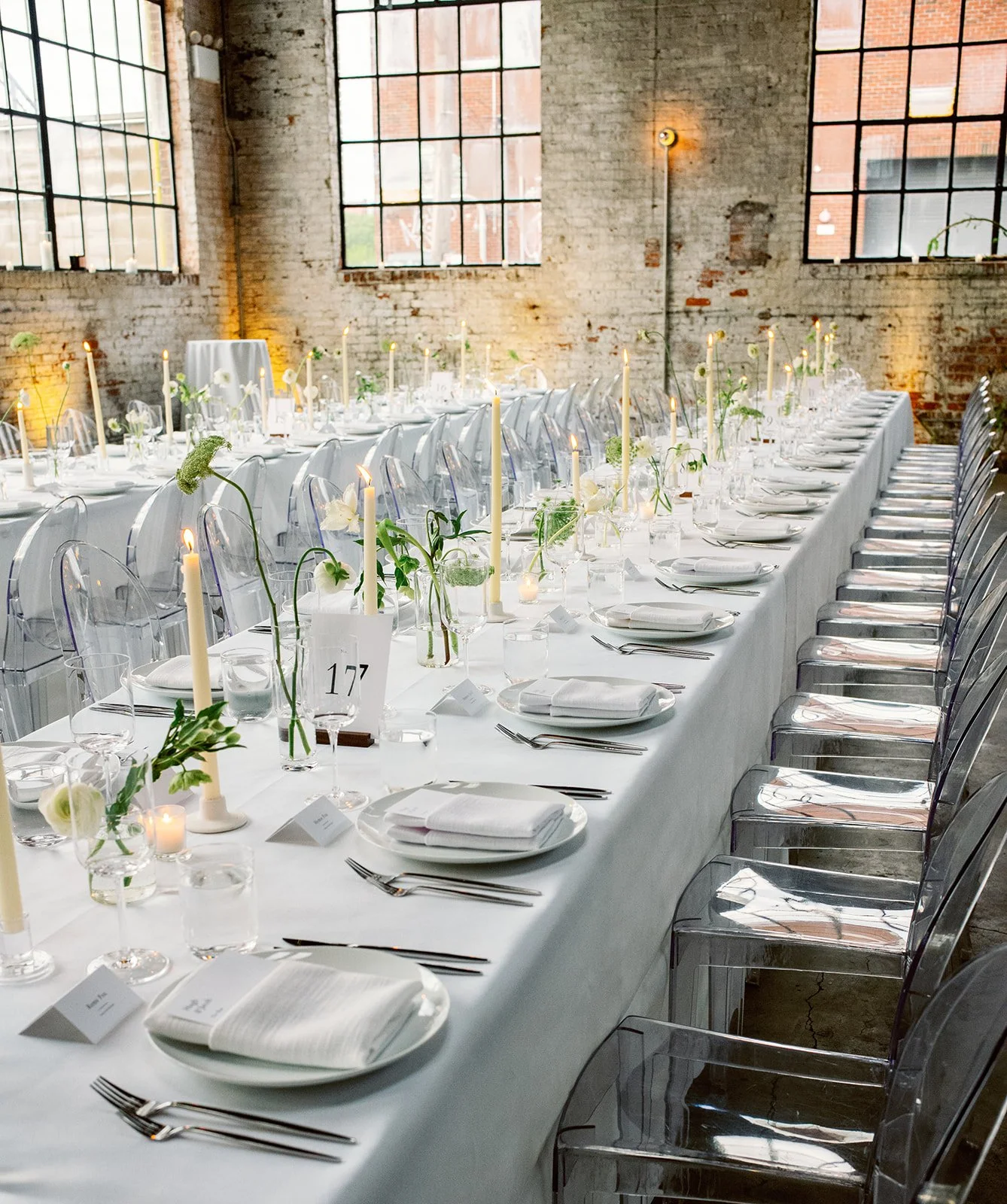 A long banquet table set for a formal event in an industrial-style venue with exposed brick walls and large windows, decorated with white tablecloths, white napkins, clear glassware, candles, and minimalist floral arrangements.
