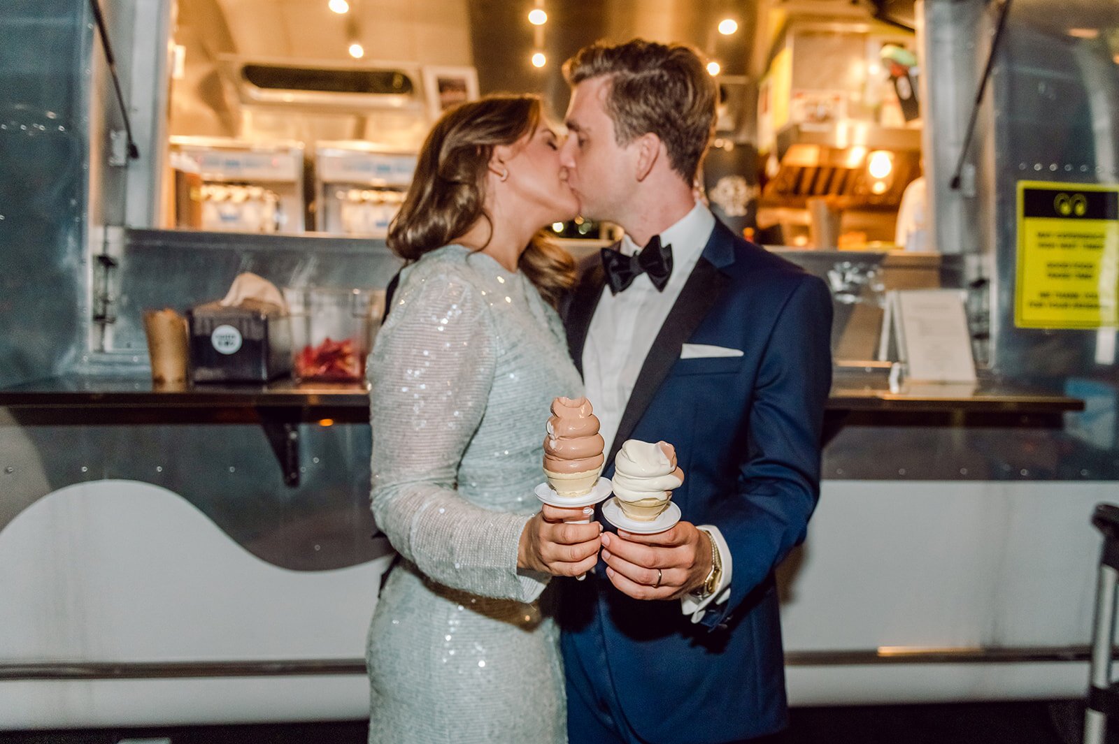 A couple in formal attire sharing a kiss while holding ice cream cones in front of a food truck.