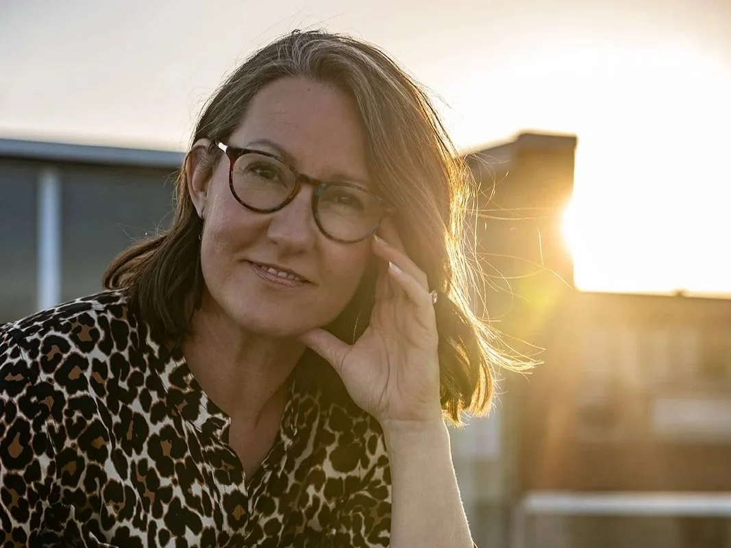A woman with shoulder-length brown hair wearing glasses and a leopard print top, smiling while touching her face, with sunlight in the background.