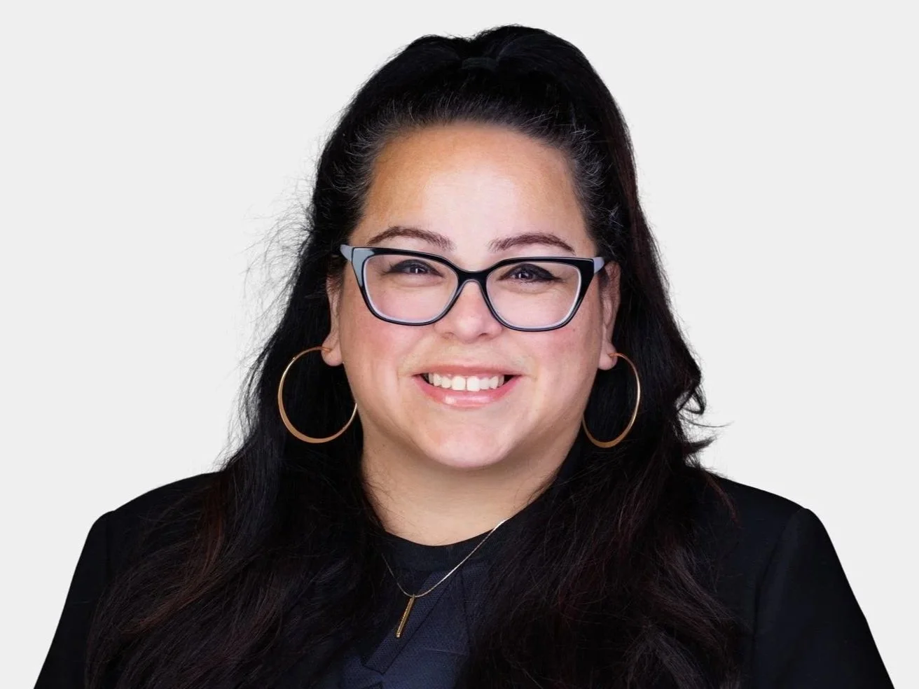 A woman with long dark hair, wearing glasses, large hoop earrings, a black blazer, and a necklace, smiling against a plain light background.