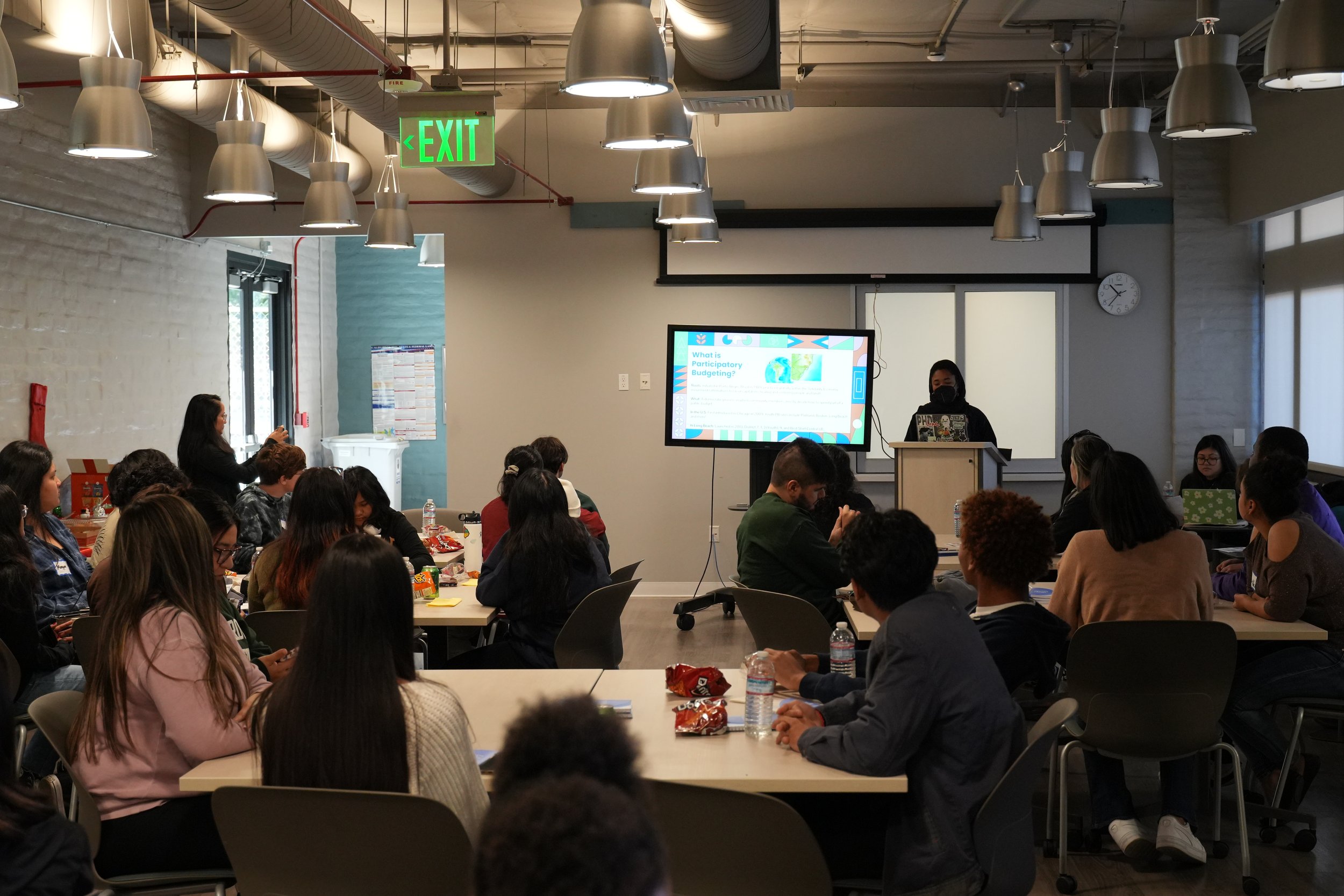 j. nyla facilitates an orientation on communty participatory budgeting for the Youth Power Participatory Budgeting system in the City of Long Beach. They stand masked at a podium while an introductory video plays.