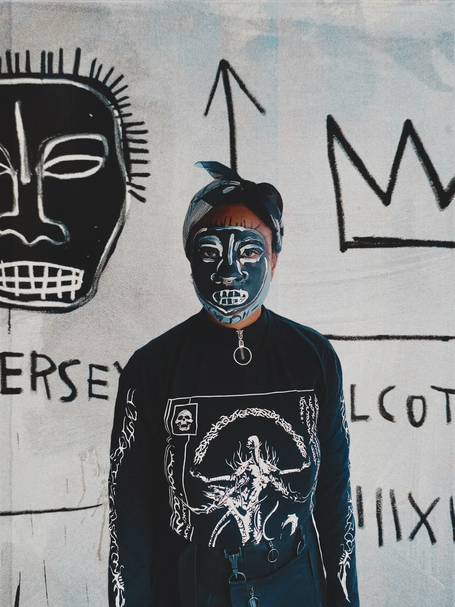 brown person wearing gothic clothing against a blown up basquiat painting, with a piece of the painting painted on their face
