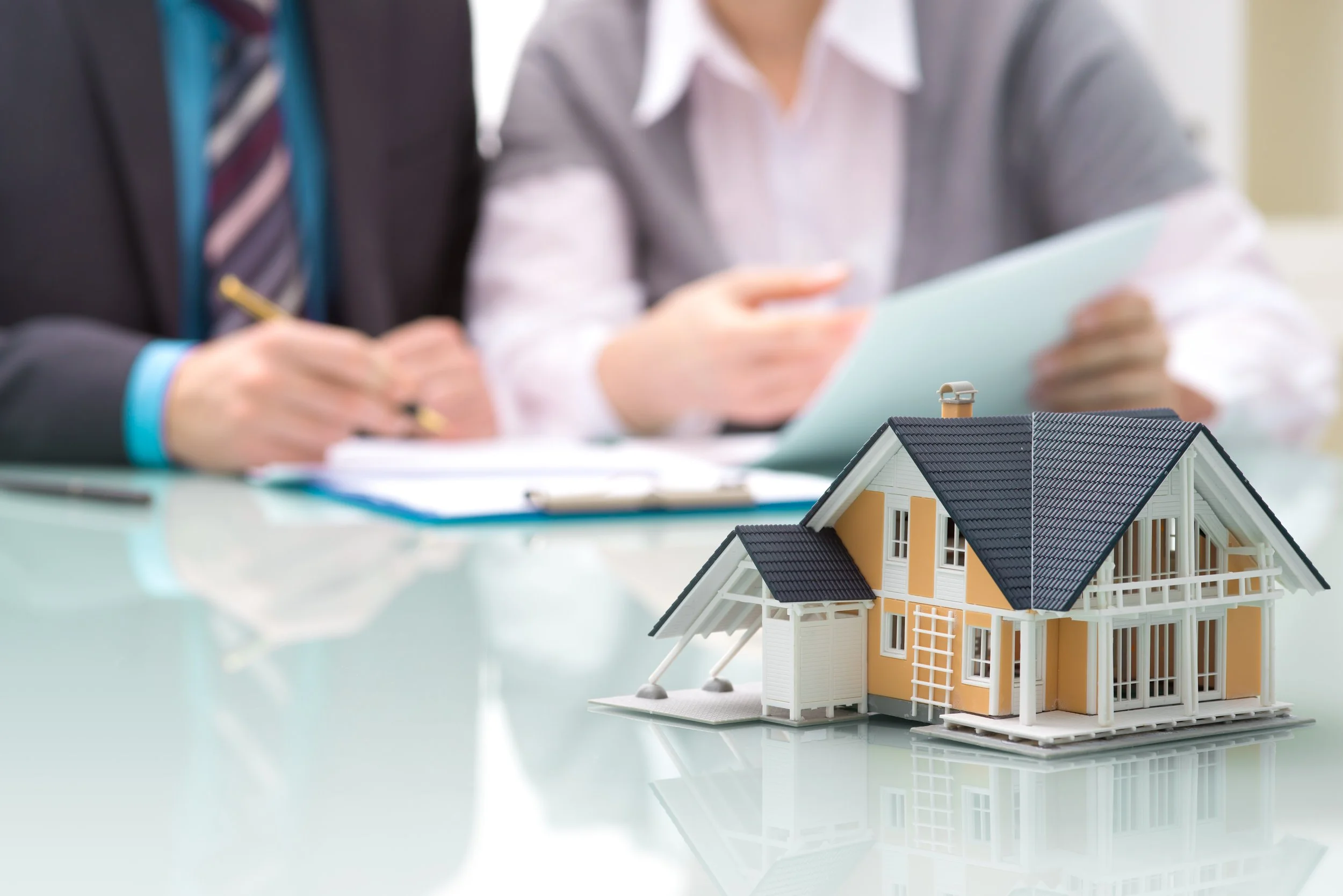 Two people signing papers at a desk beside a small model house