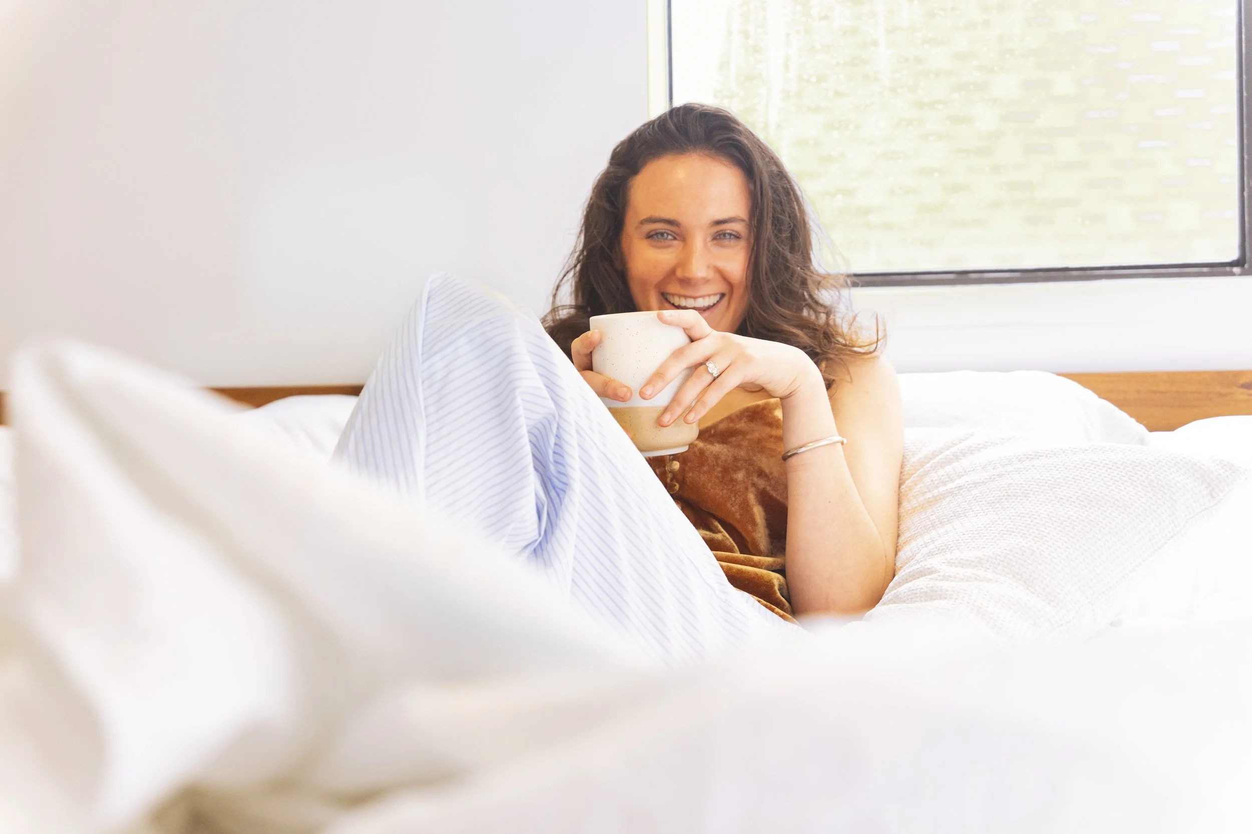 A woman with curly brown hair smiling and holding a beige mug while sitting in bed near a window.