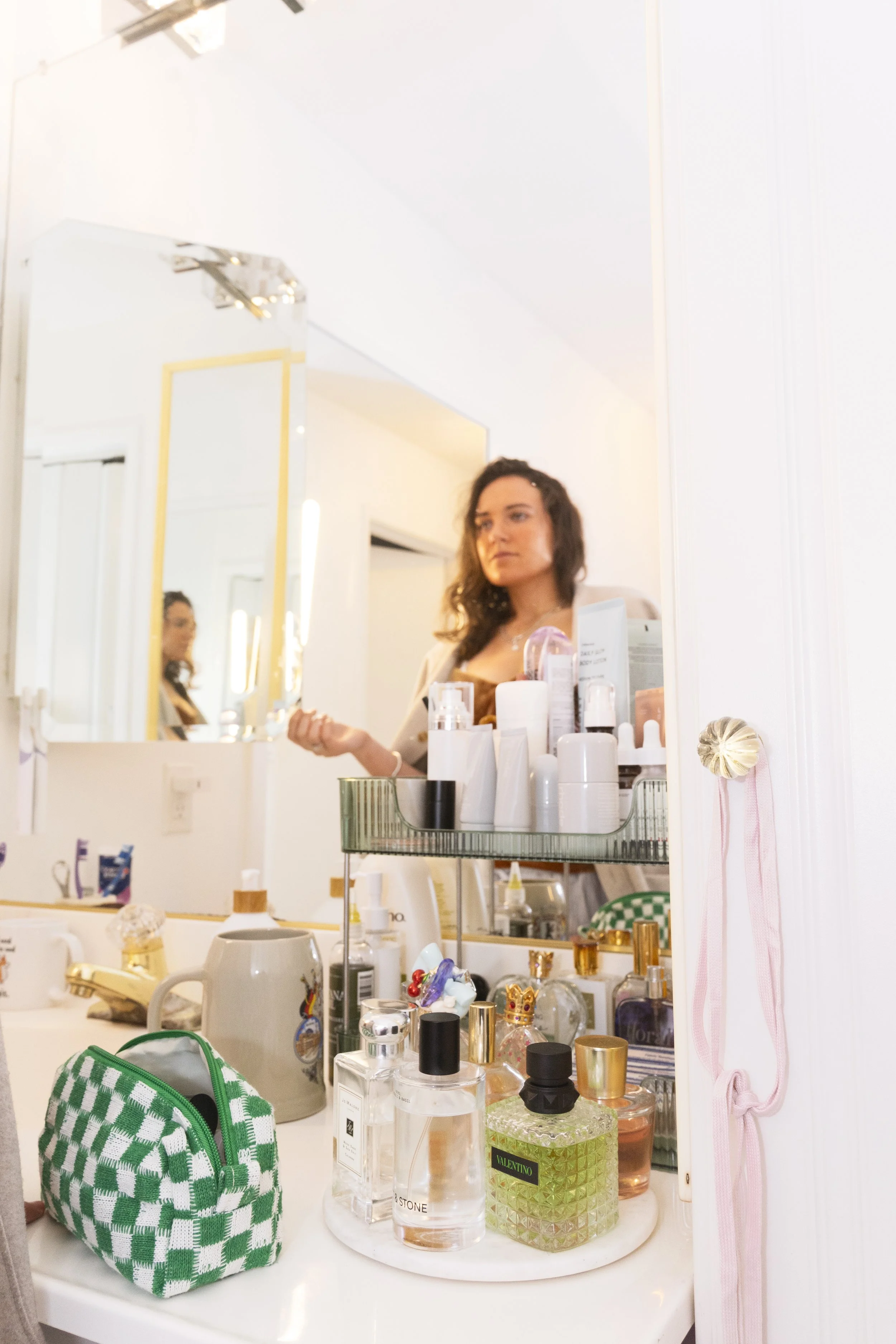A young woman seen through a mirror in a bathroom, holding a spoon, surrounded by toiletries and perfume bottles on the countertop.
