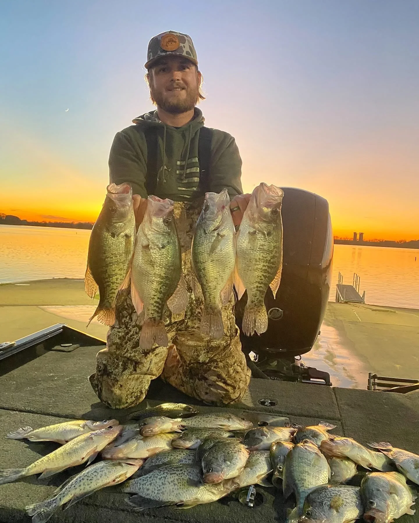 A man kneeling on a boat deck at sunset holding four large fish, with additional fish laid out in front of him on the deck, near a body of water with two industrial structures in the distance.