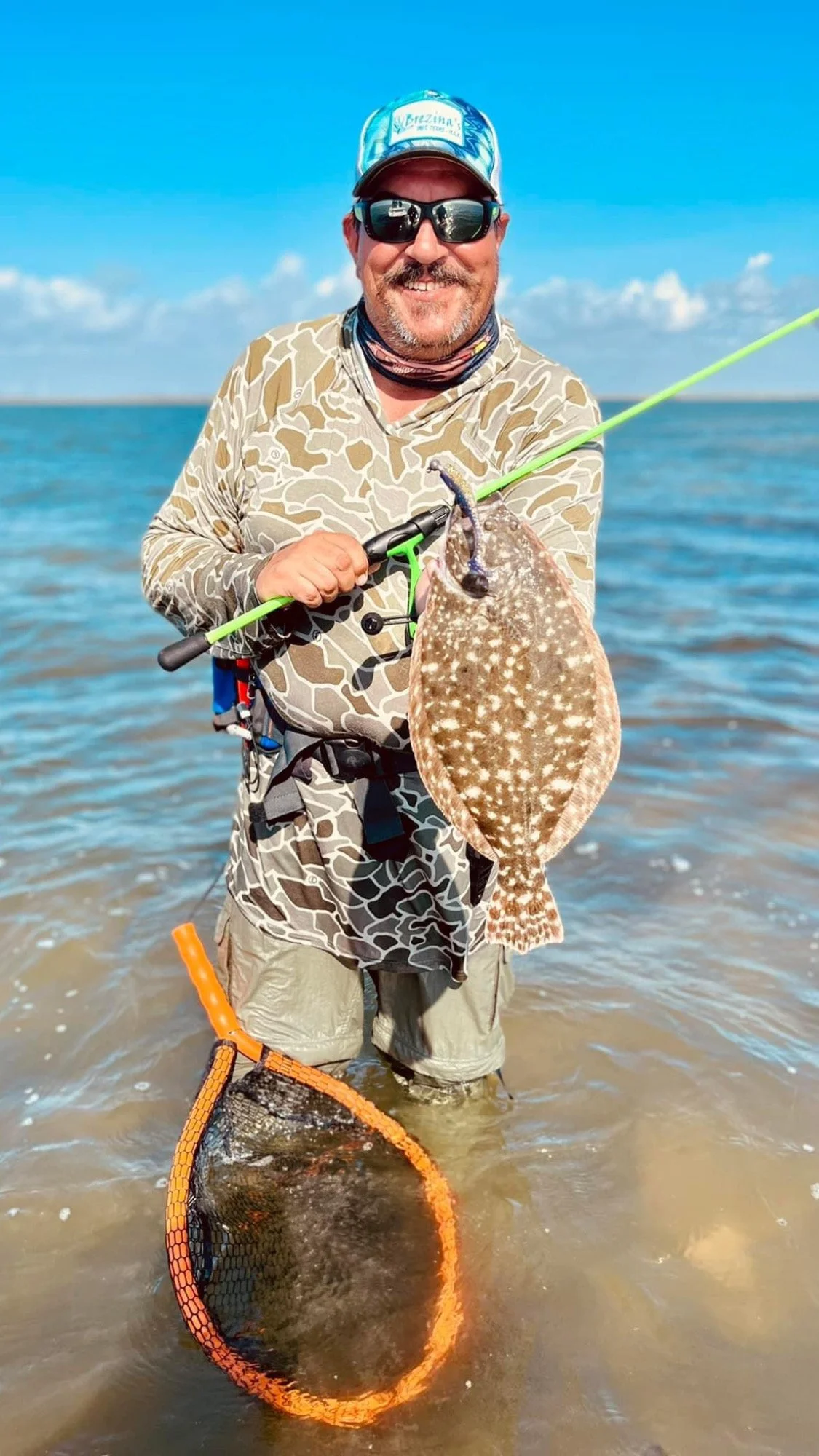 Man holding a flounder caught while fishing in shallow water, wearing camo shirt and cap, with a fishing rod and net.
