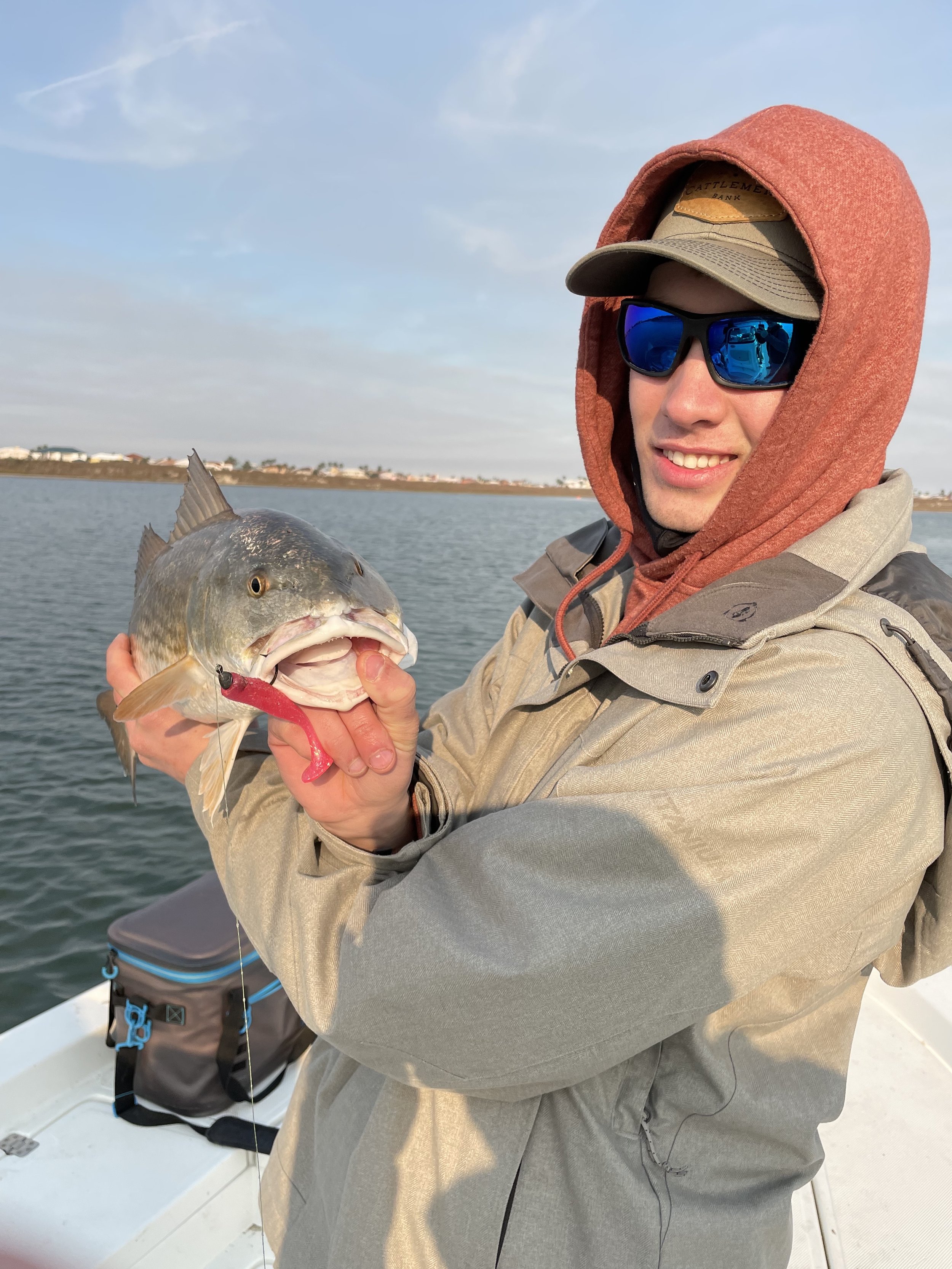 Person holding a fish on a boat, wearing sunglasses, a brown hoodie, and a gray jacket.