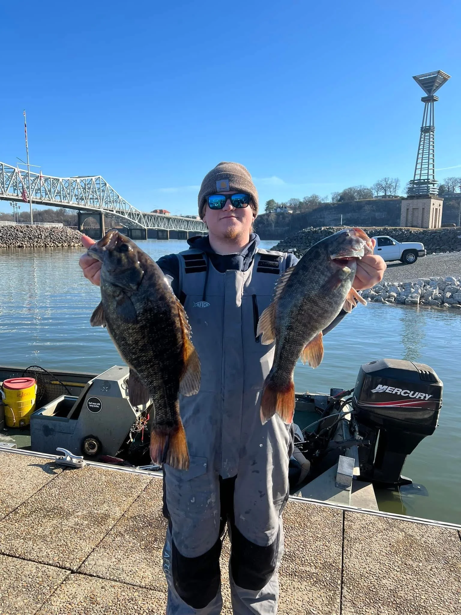 A man wearing sunglasses, a gray jacket, and a beanie hat is standing by a body of water, holding two large fish, one in each hand. There is a boat with a Mercury motor behind him and a bridge and water tower in the background under a clear blue sky.