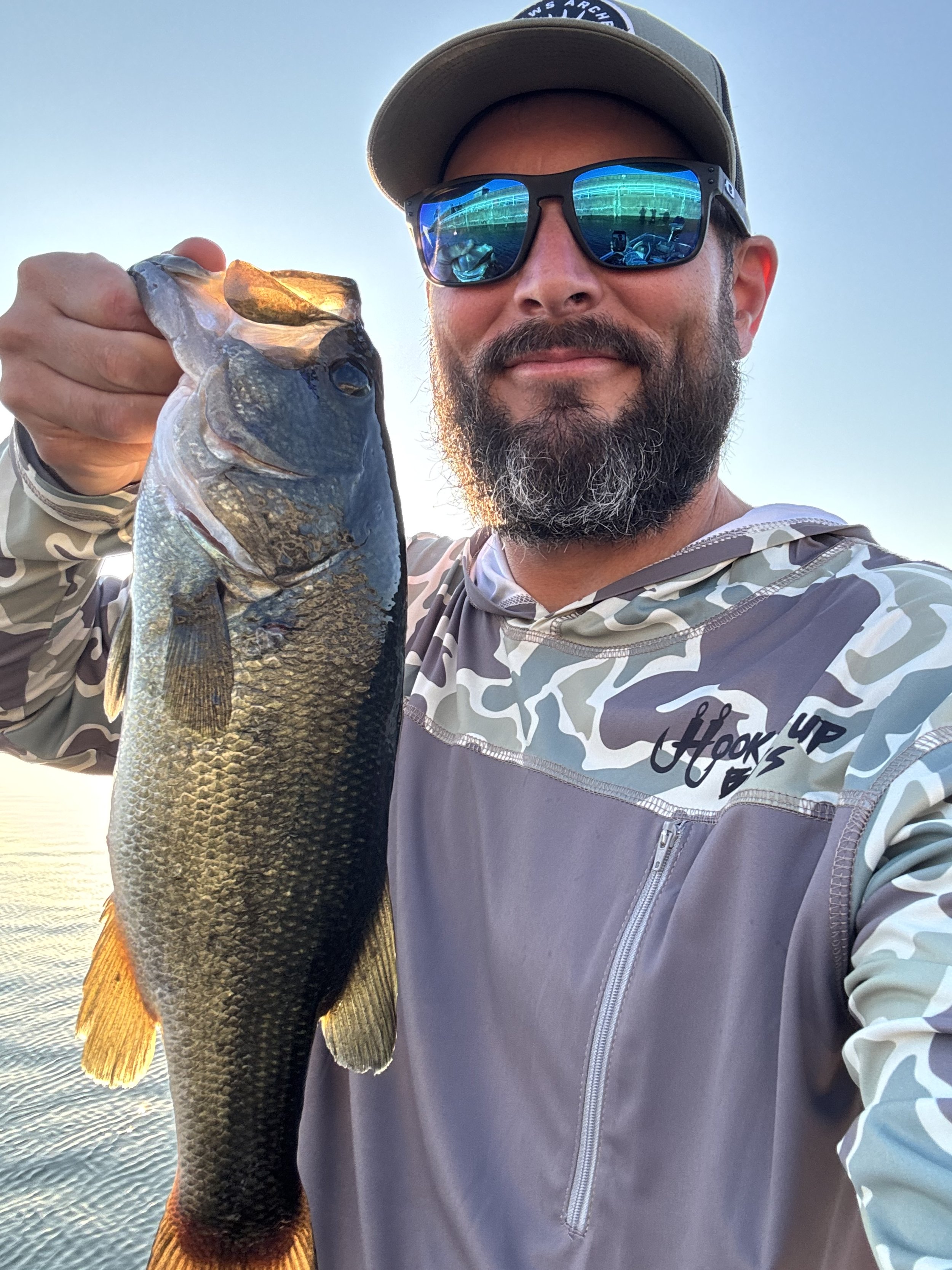 A man with a beard wearing sunglasses, a cap, and a camo-patterned jacket holding a large fish in his hand during sunset or sunrise over a body of water.