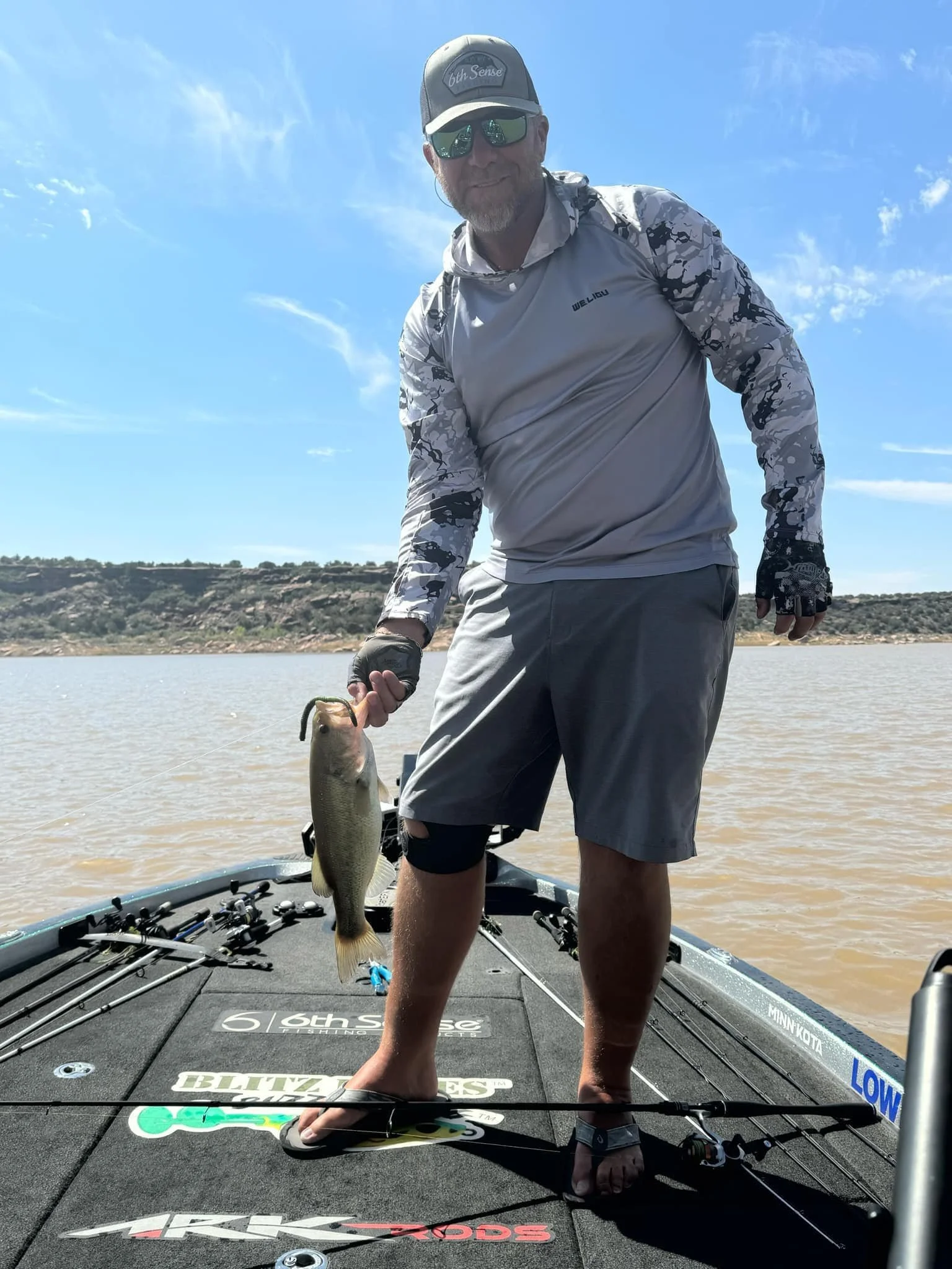Man on a boat holding a fish under a clear blue sky, wearing a cap, sunglasses, long-sleeved shirt, and shorts.