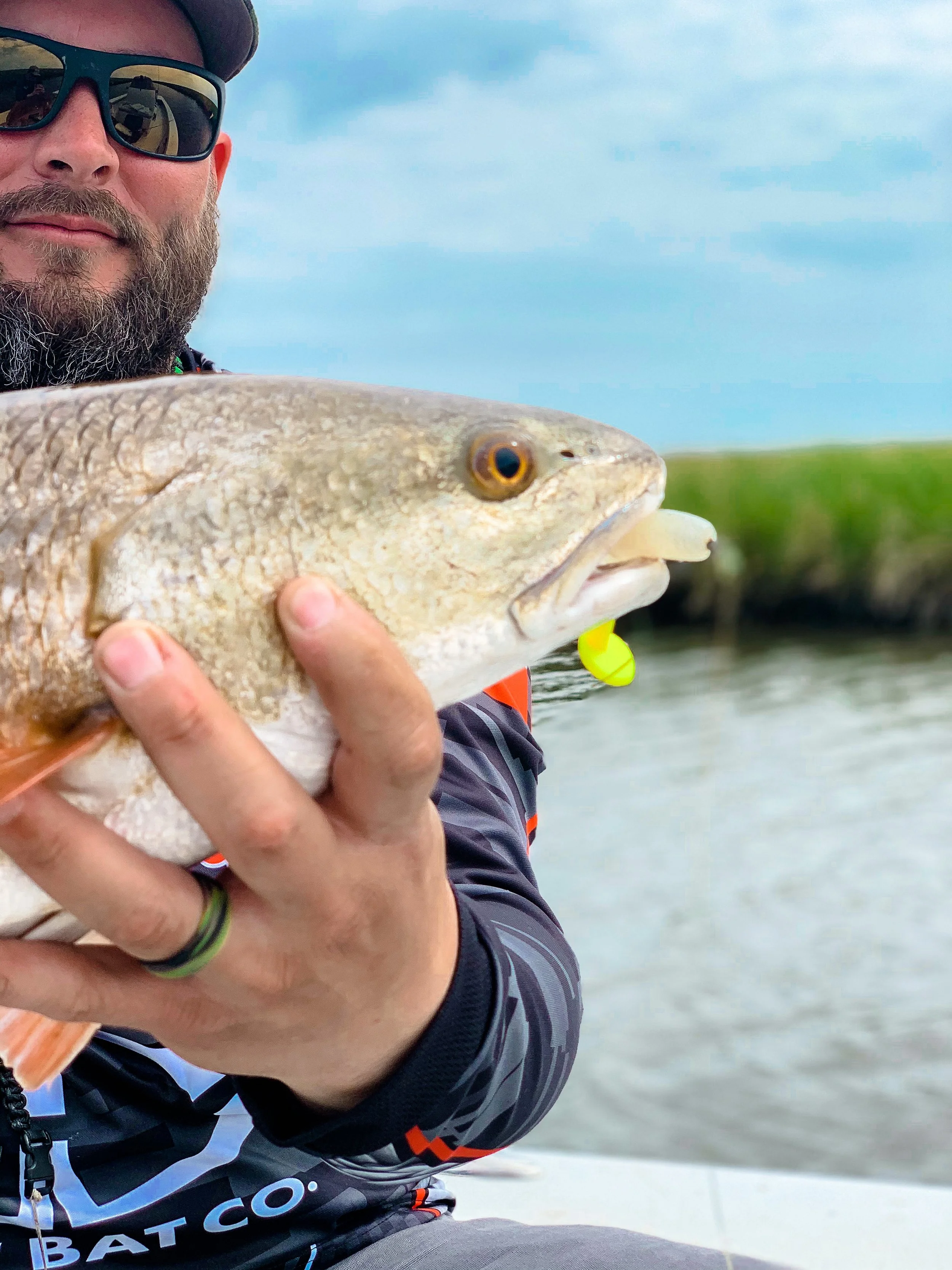 Man holding a fish with a lure in its mouth by a body of water, cloudy sky in the background.