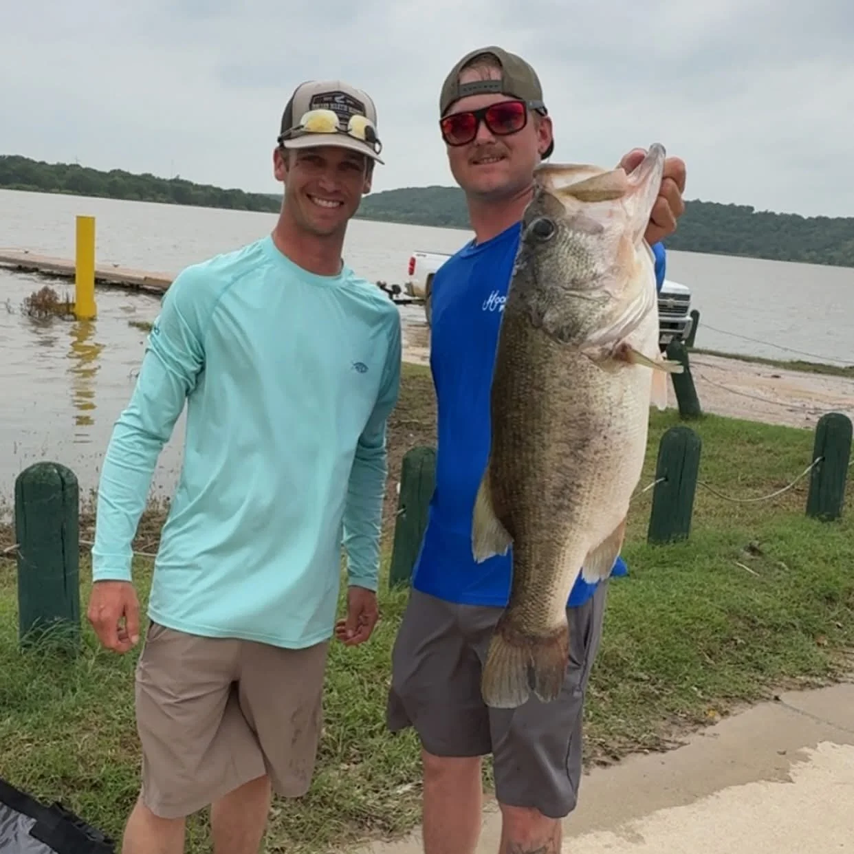 Two smiling men, one holding a large fish, standing near a body of water with a dock and greenery in the background.