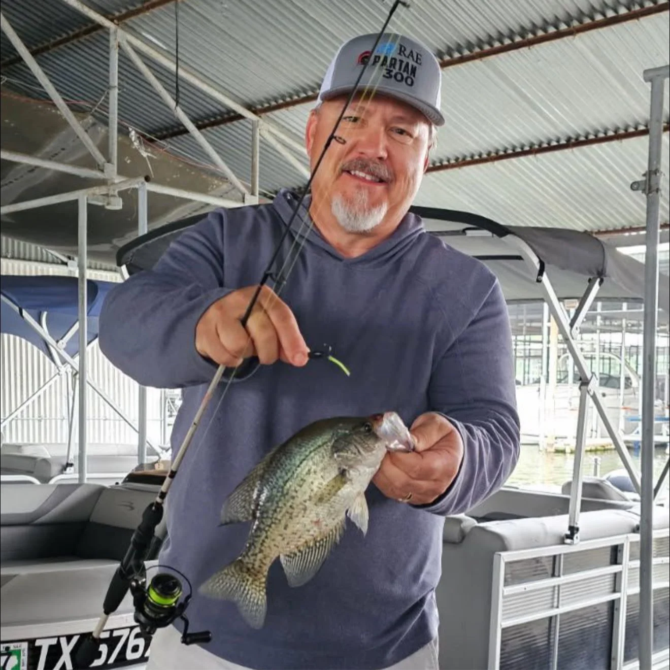 A man in a gray hoodie and cap holding a fishing rod and a large fish in a marina with boats and a covered dock.