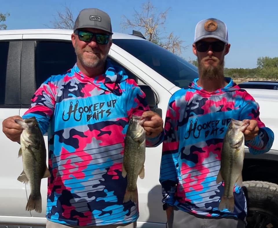 Two men wearing matching camouflage-patterned fishing jerseys with 'Hooked Up Baits' written on them, holding up fishing catches in front of a white vehicle outdoors on a sunny day.