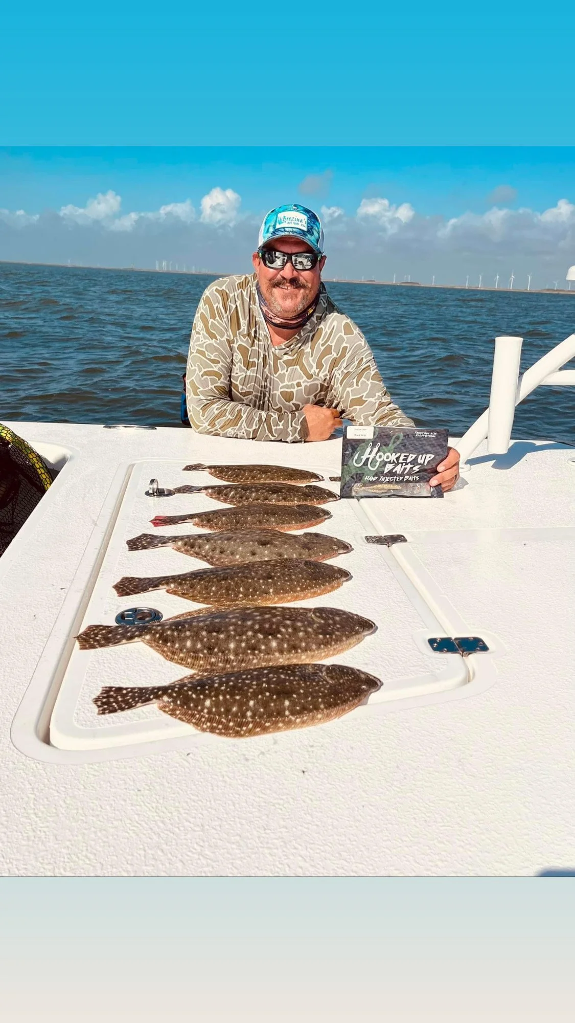 Man on a boat displaying several flatfish on a cutting board, with open water in the background, under a clear blue sky.