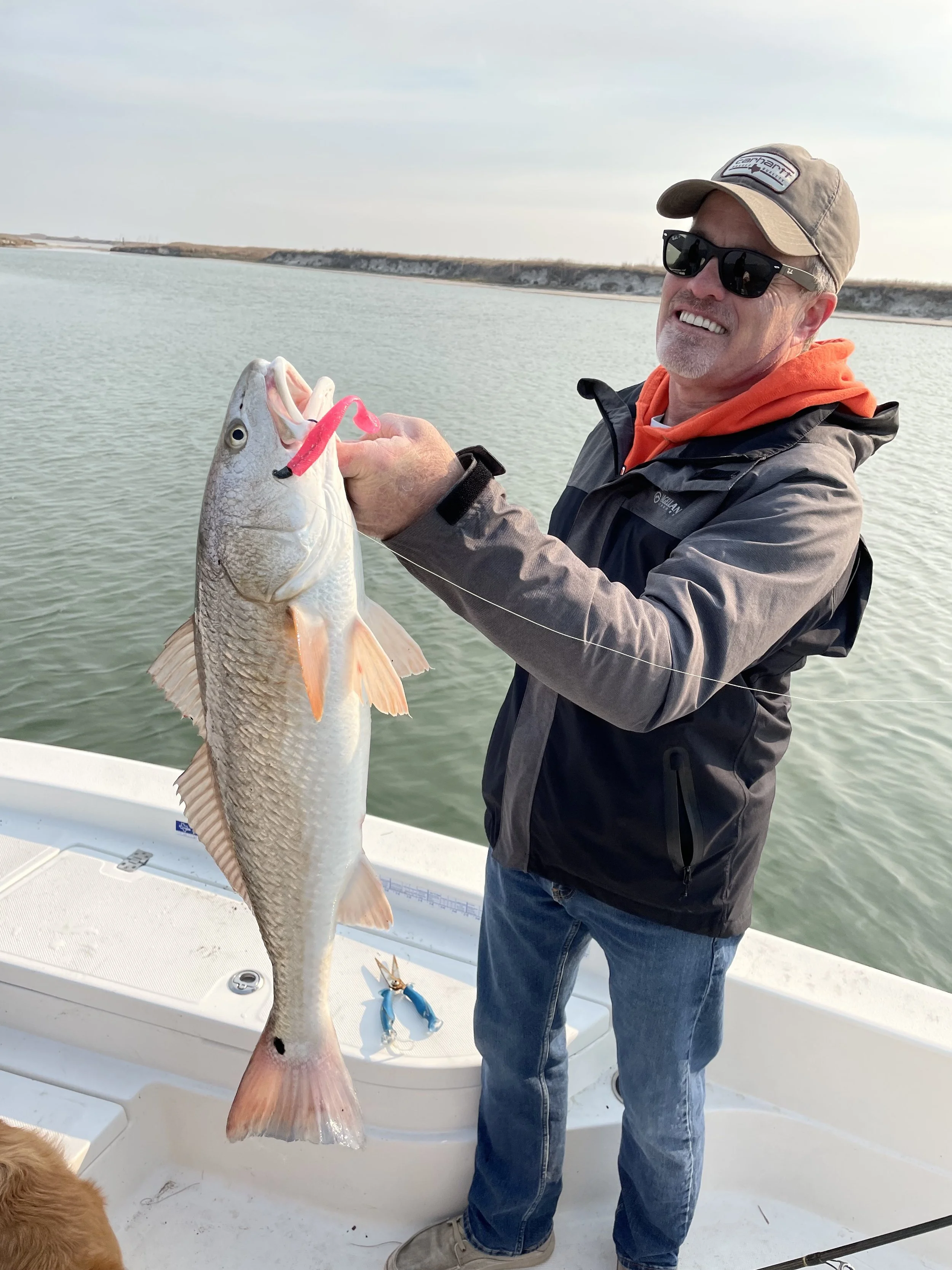 Man holding a large fish on a boat, wearing a cap and sunglasses, with water in the background.