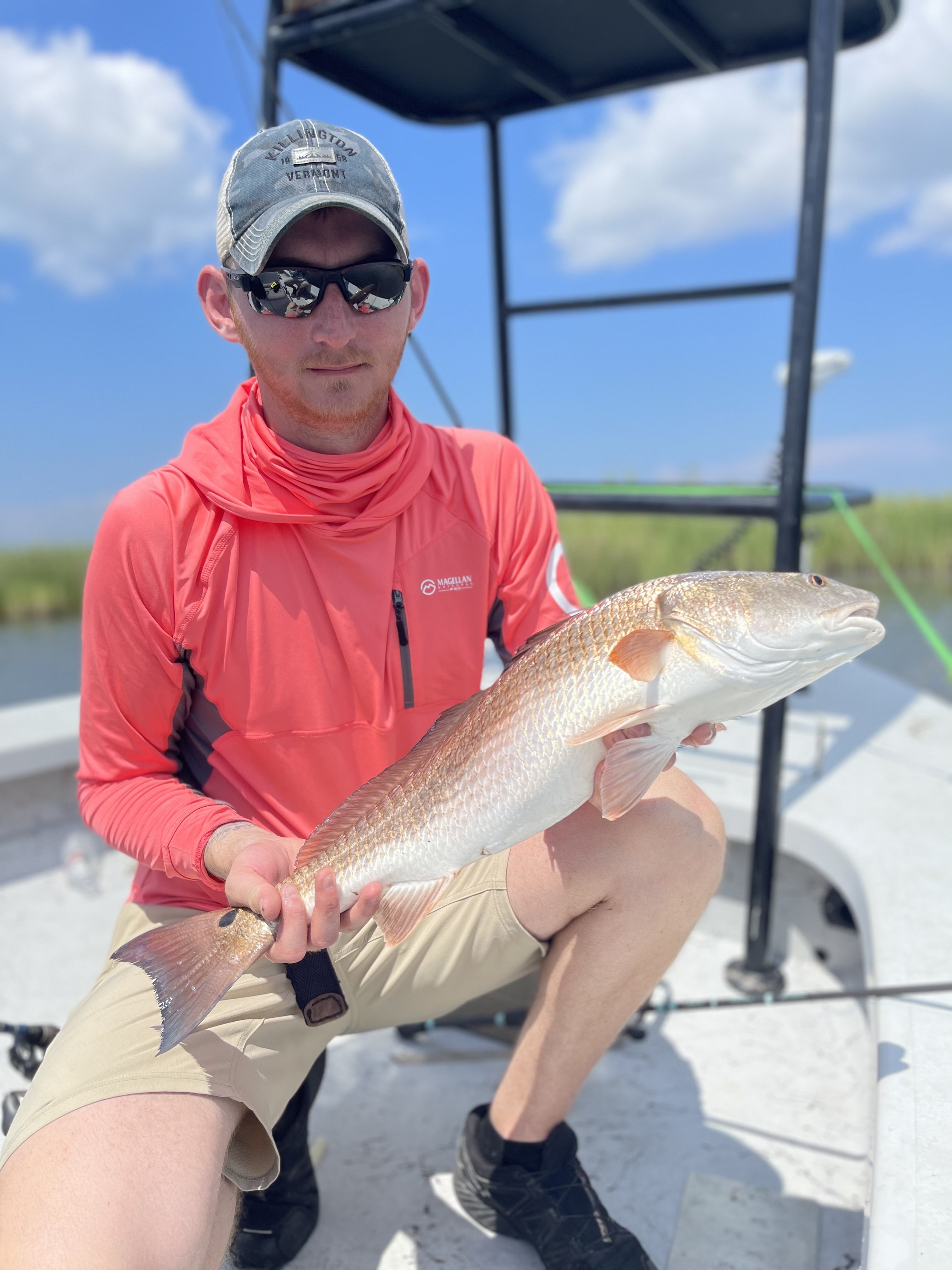 A person wearing a cap and sunglasses holds a fish while kneeling on a boat under a blue sky.