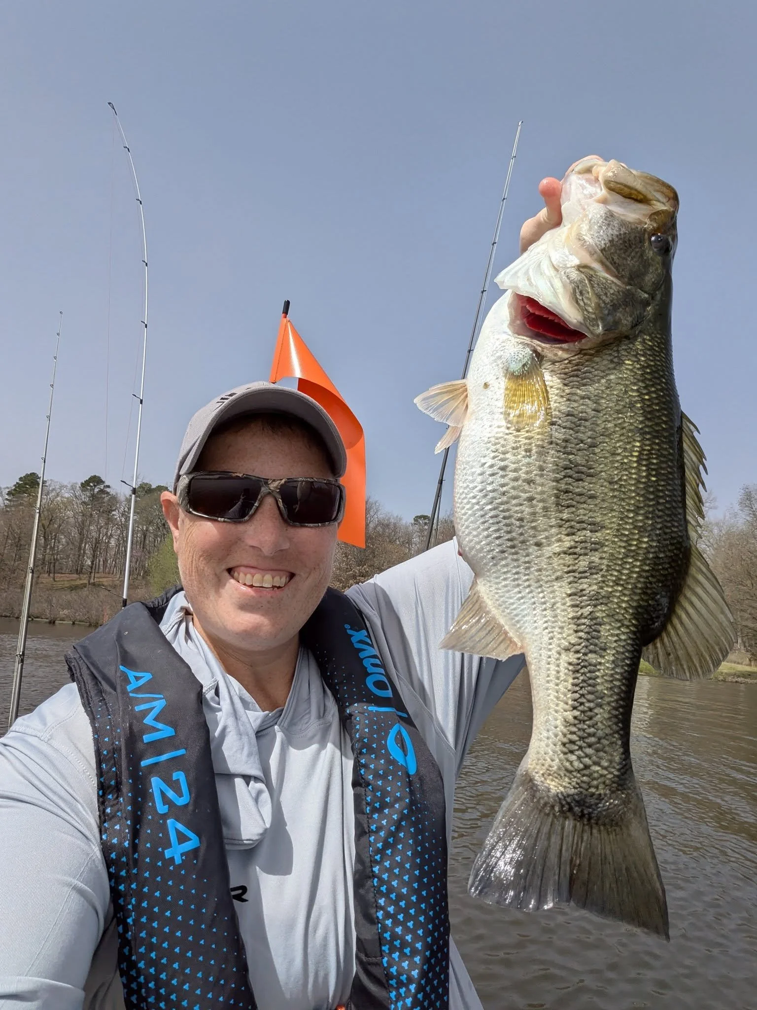 A smiling man wearing sunglasses, a gray cap, and a life vest holding a large bass fish on a boat in a river with trees in the background.
