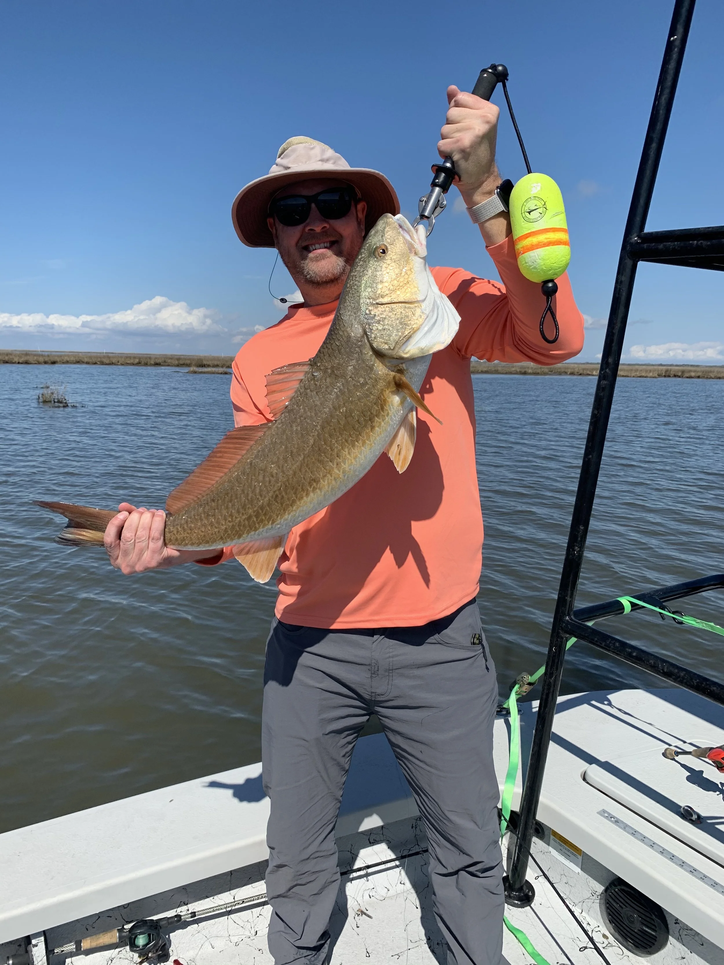 Person holding a large fish on a boat with water and grass in the background, wearing a hat and sunglasses.