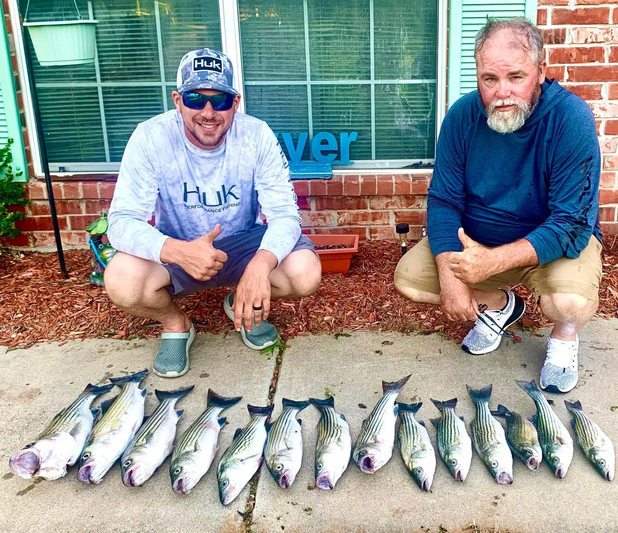 Two men crouching on either side of a row of striped bass fish laid out on the ground, posing with thumbs up. They are in front of a window with brick walls and some plants nearby.