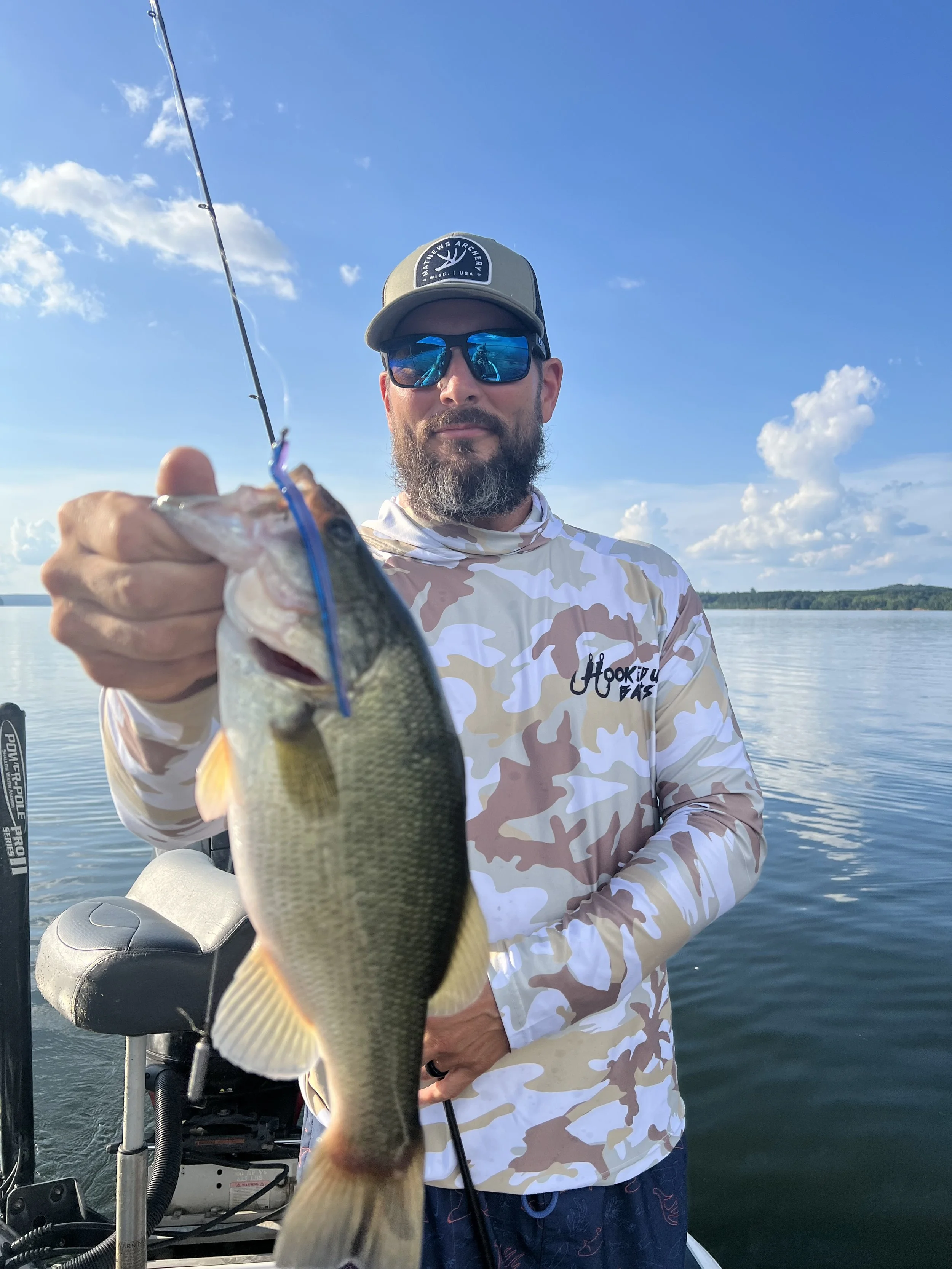 Man holding a fish he caught on a fishing trip, standing on a boat on a lake with blue sky and clouds in the background.