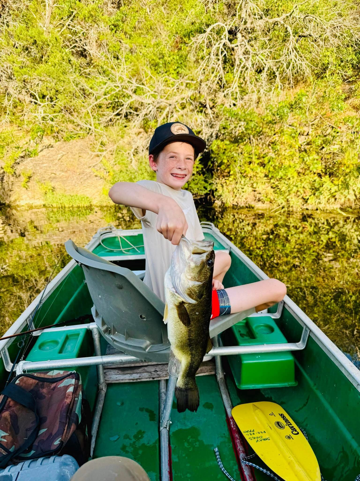 A smiling young boy sitting in a small boat on a calm river, holding up a large fish he caught, with green trees and reflections on the water in the background.