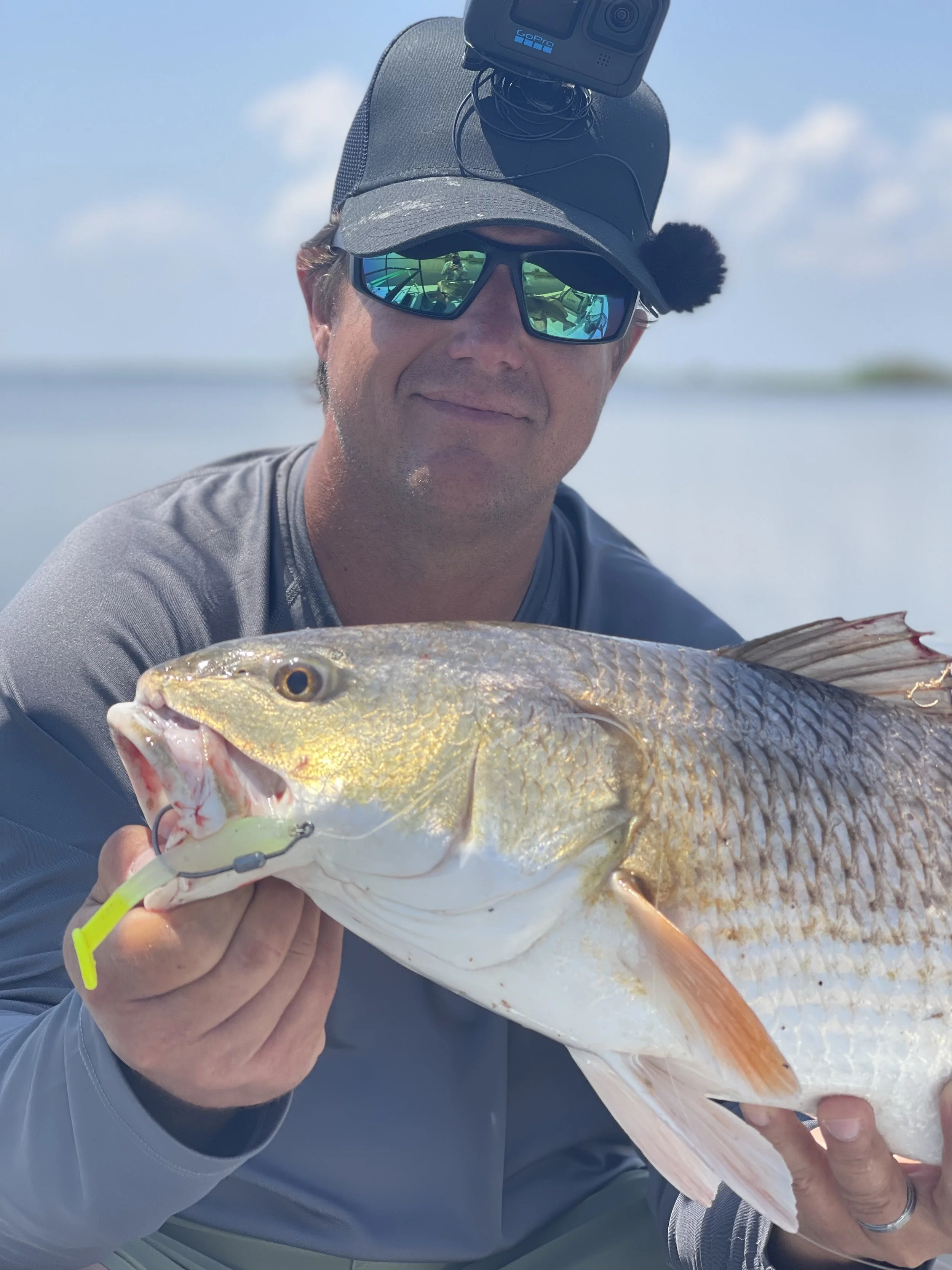 Man holding a large fish with a lure in its mouth, wearing a cap with a mounted camera, and sunglasses, against a clear blue sky.