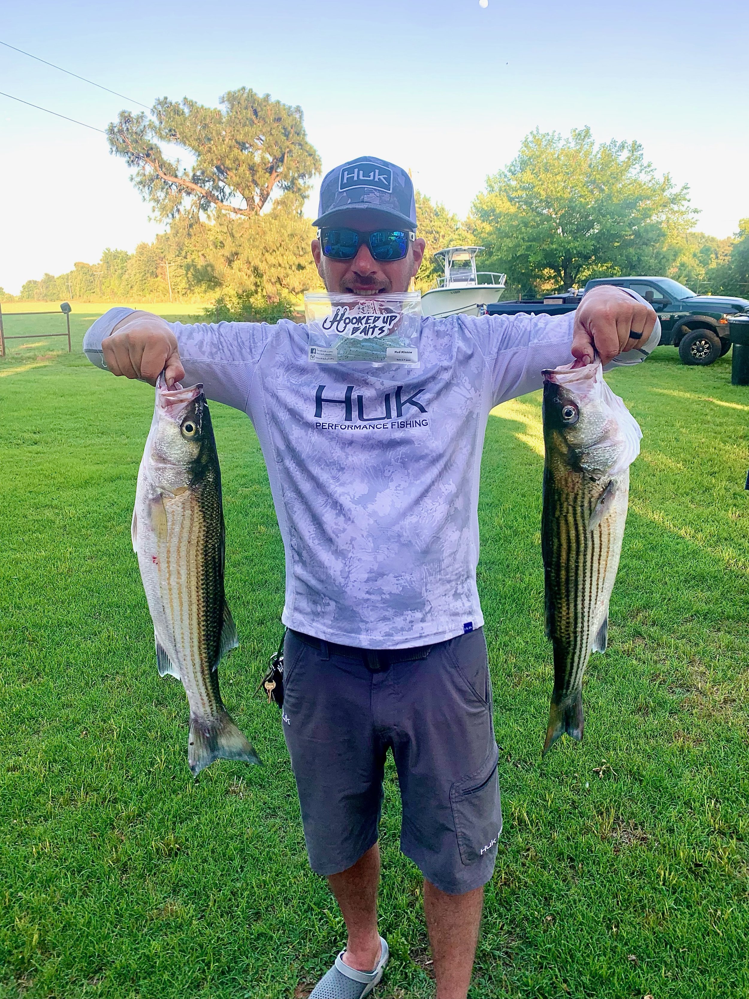 Person holding two striped bass fish, standing on grass, wearing a "Huk" fishing shirt and hat. A boat and truck are visible in the background.
