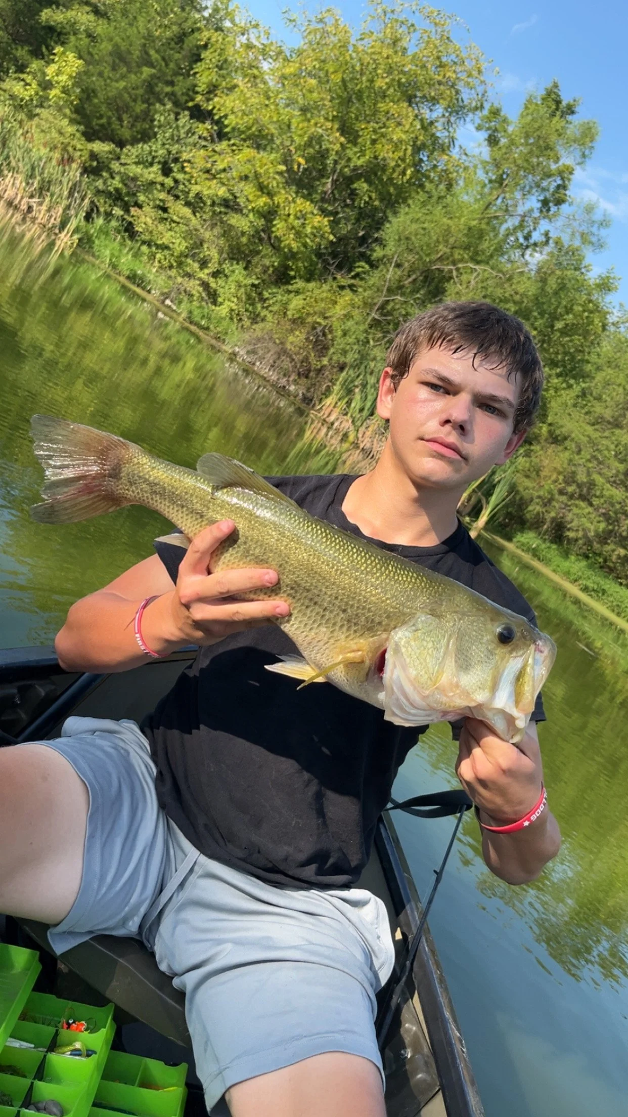 A young man sitting in a boat on a river, holding a large fish he caught, with lush green trees and blue sky in the background.