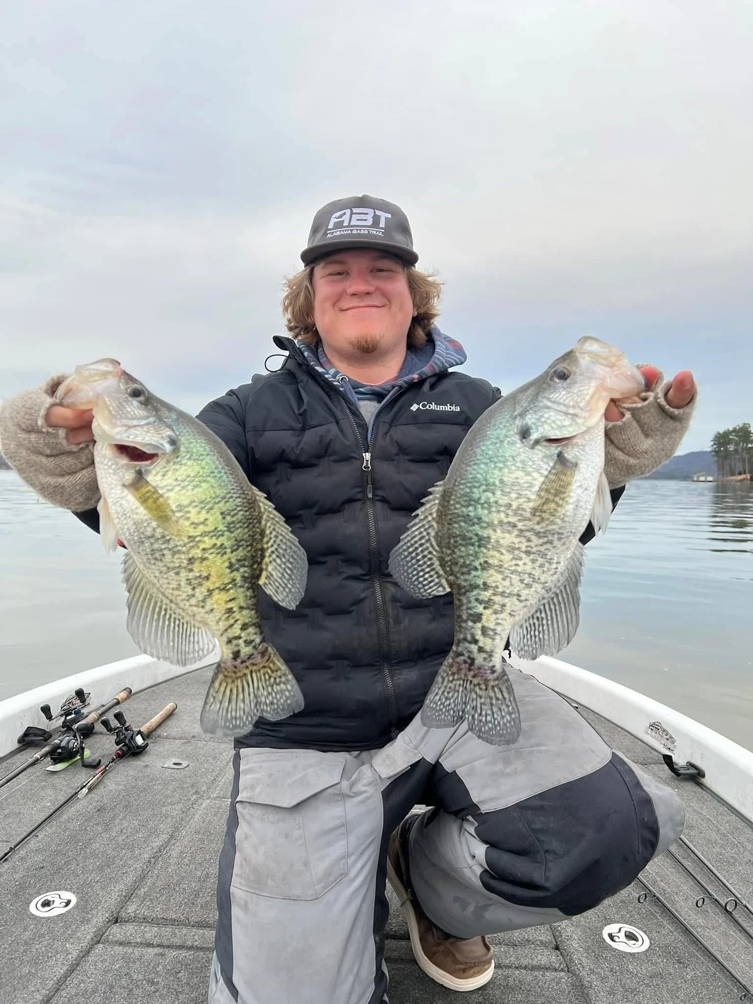 A person holding two large fish on a boat on a calm lake with a cloudy sky.