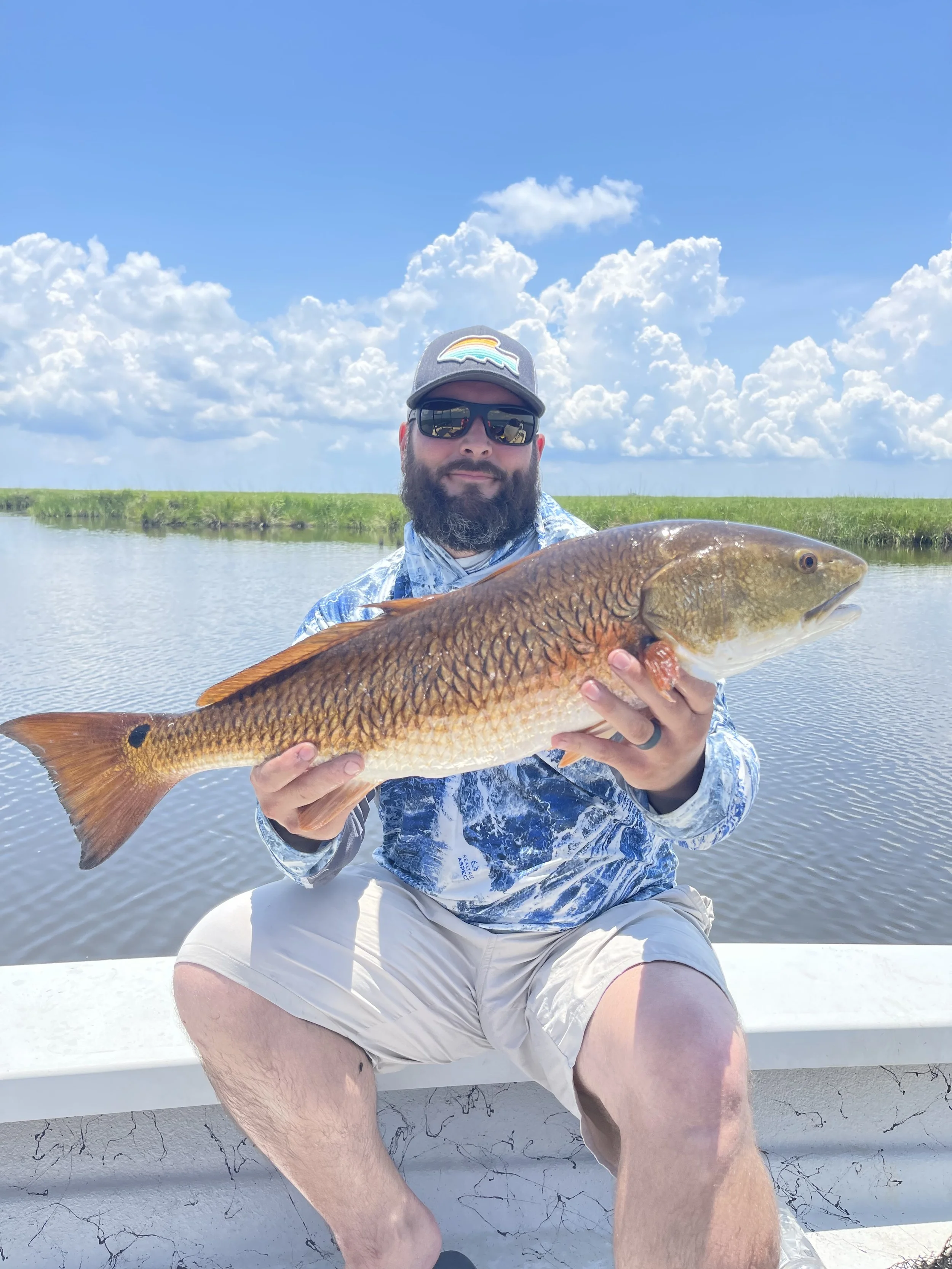 A person wearing sunglasses and a cap holding a large fish on a boat, with a backdrop of water and cloudy sky.