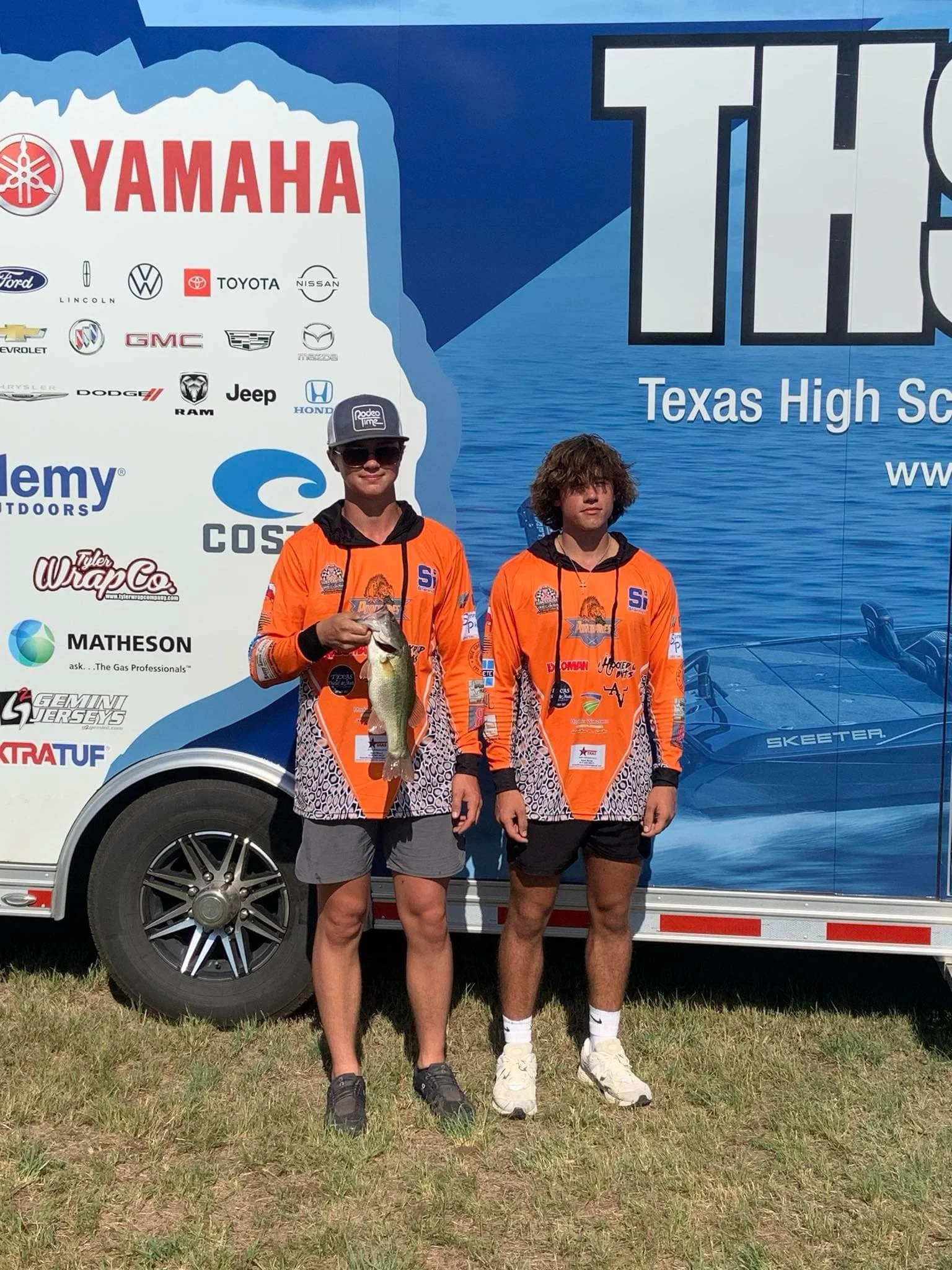 Two young men in orange fishing jerseys holding a fish in front of a trailer with various brand logos, including Yamaha and Texas High School Bass Association.