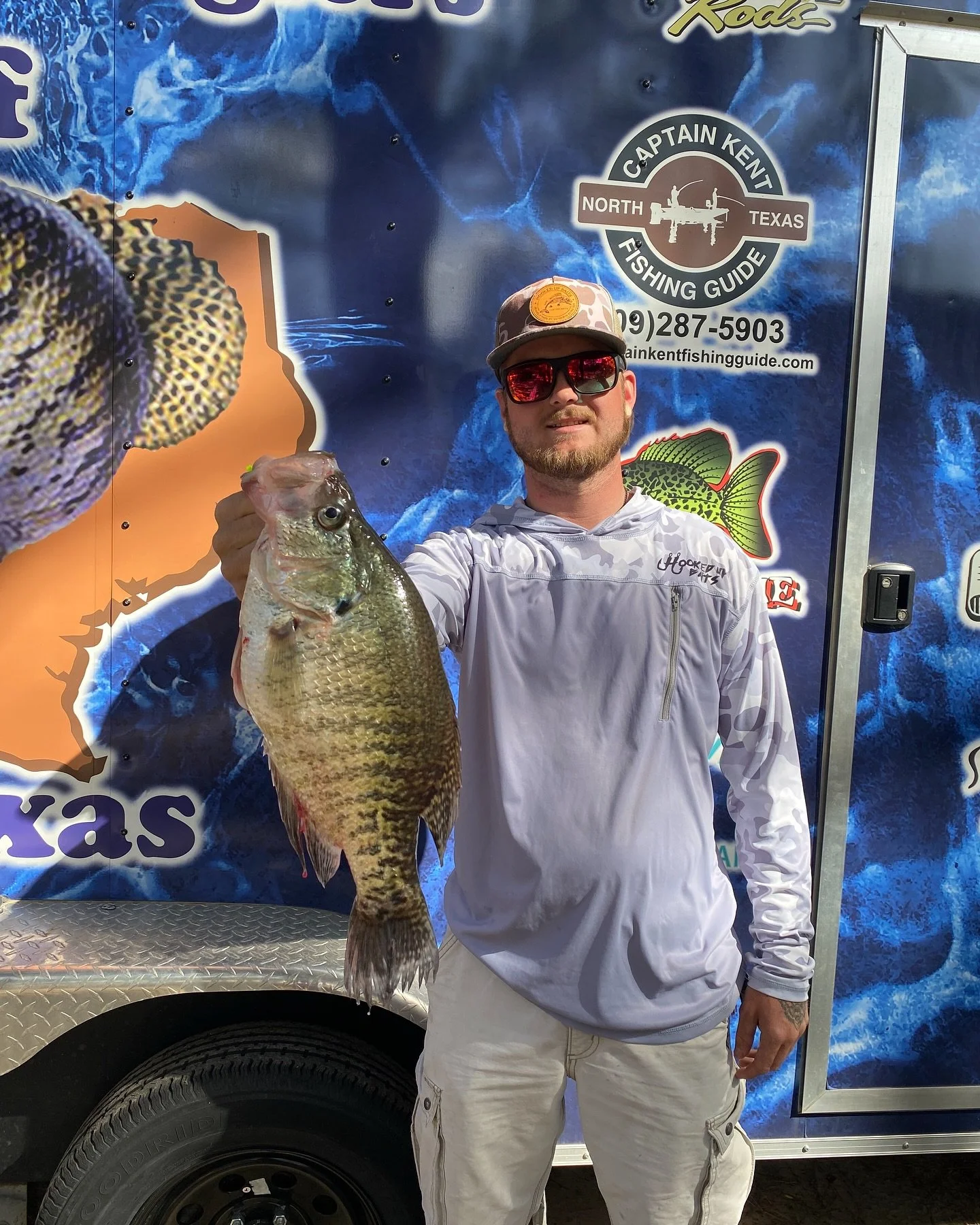 A man wearing sunglasses, a cap, and a white long-sleeve shirt holding a large fish in front of a blue fishing guide's trailer. The trailer has graphics and contact information for Captain Kent, a fishing guide in North Texas.