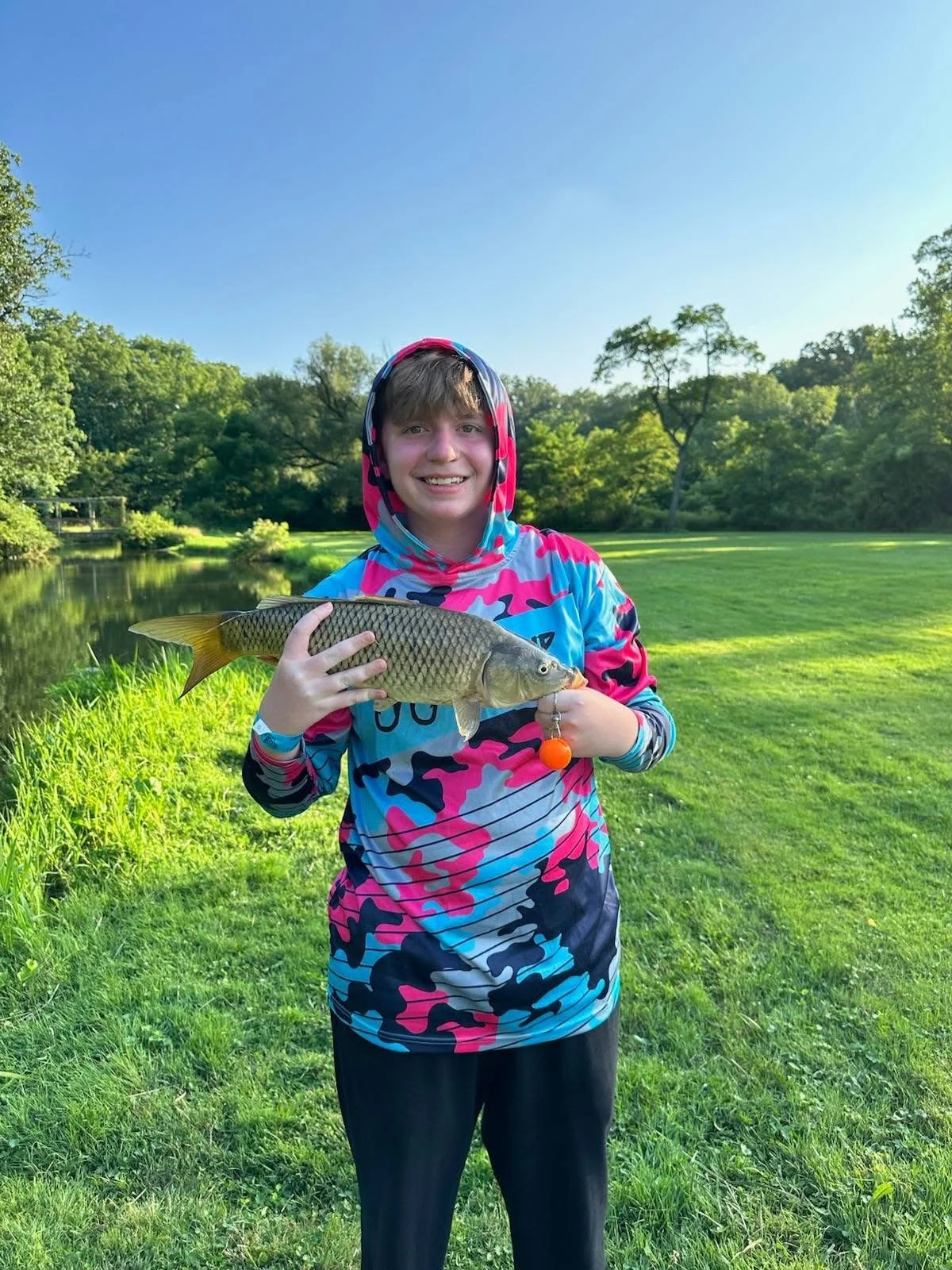 A person in colorful camouflage hoodie holding a fish outdoors near a pond and grassy area, smiling at the camera.